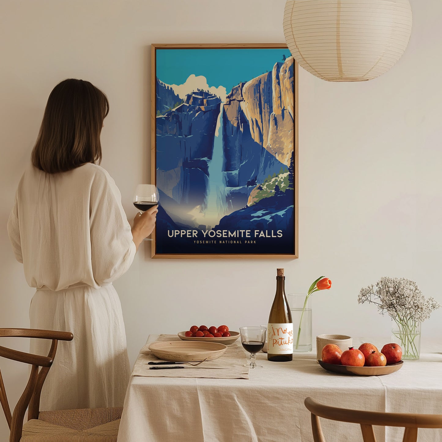 A woman in a white dress holds a wine glass, admiring a framed Upper Yosemite Falls poster on the wall. The dining table is set with fruit, wine, and flowers.
