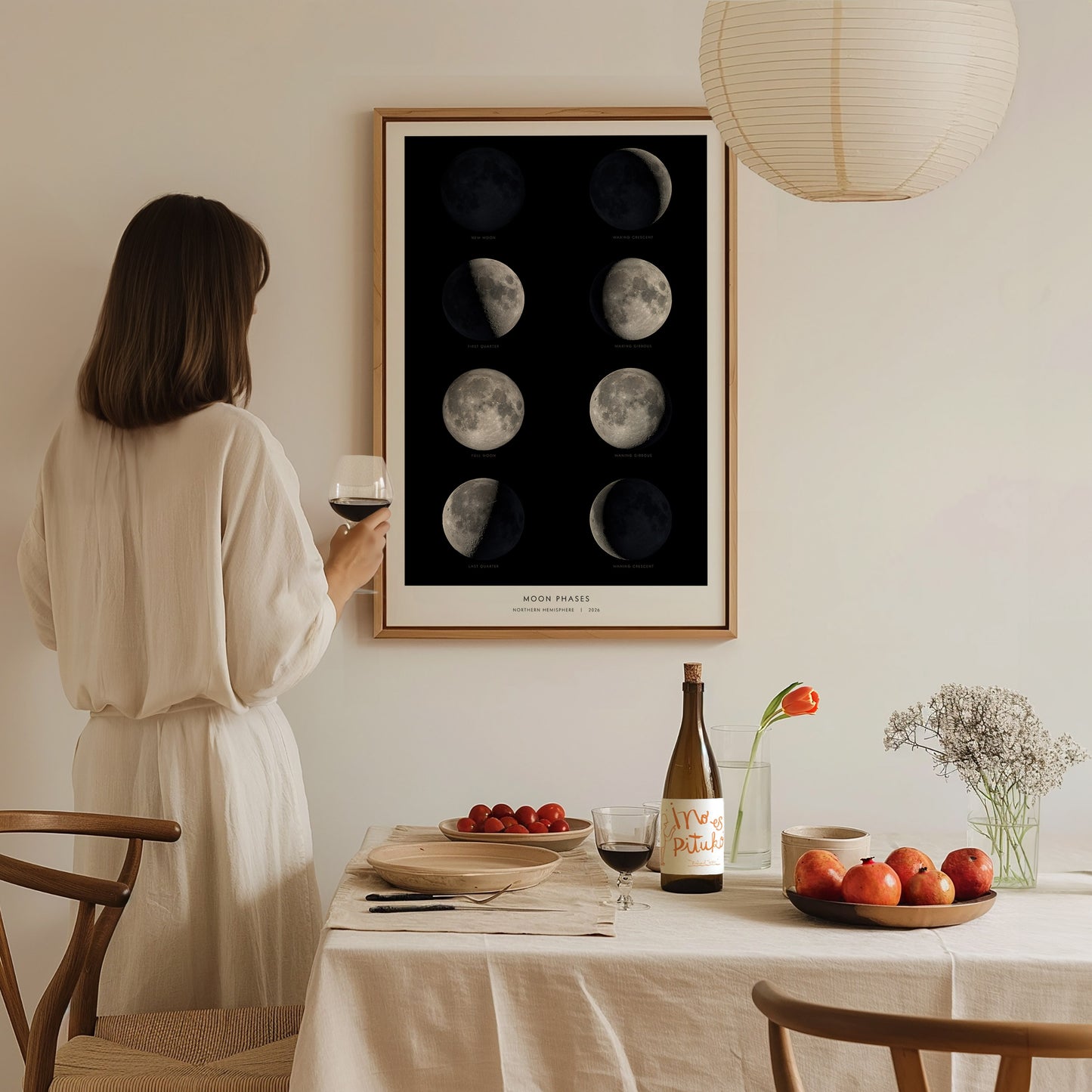 A woman in a white dress stands holding a glass of red wine, gazing at a framed moon phases poster on the wall.