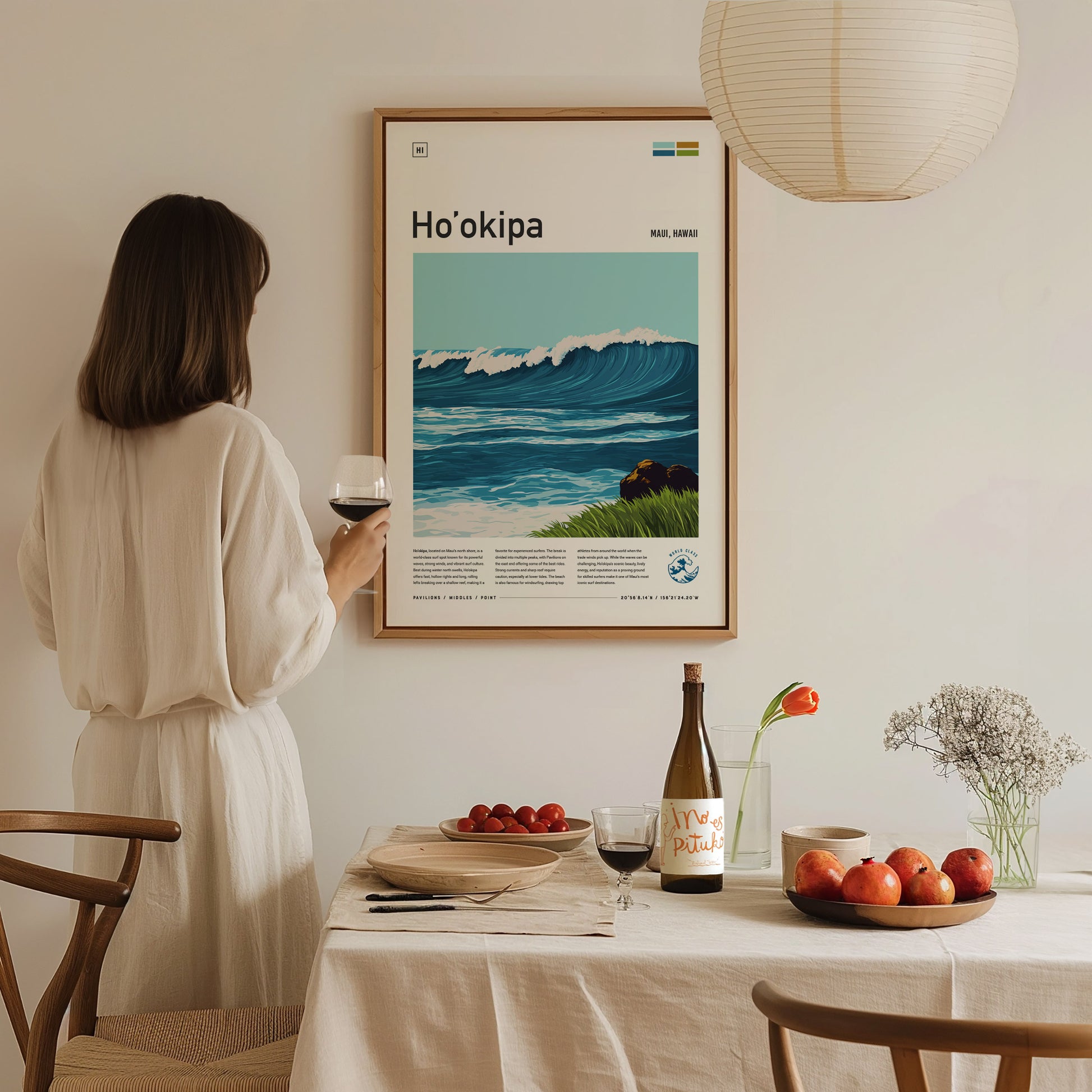 A woman in a white dress holds a glass of wine, admiring a framed Hookipa poster. A table with wine, apples, and flowers is in the foreground.