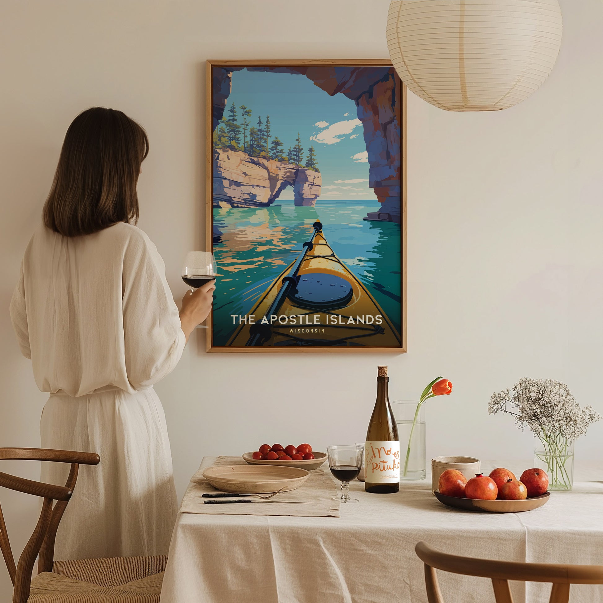 A person stands in a cozy dining area, gazing at a framed poster of the Apostle Islands, featuring a kayak gliding through serene turquoise waters surrounded by cliffs.