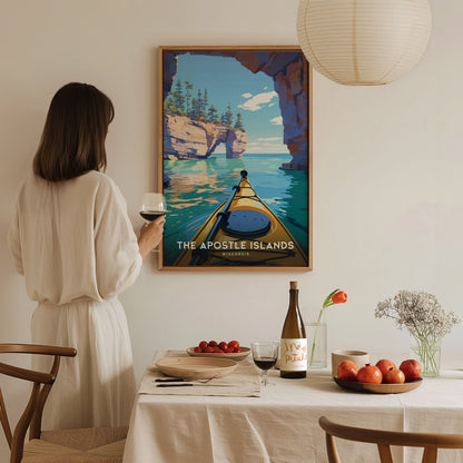 A person stands in a cozy dining area, gazing at a framed poster of the Apostle Islands, featuring a kayak gliding through serene turquoise waters surrounded by cliffs.