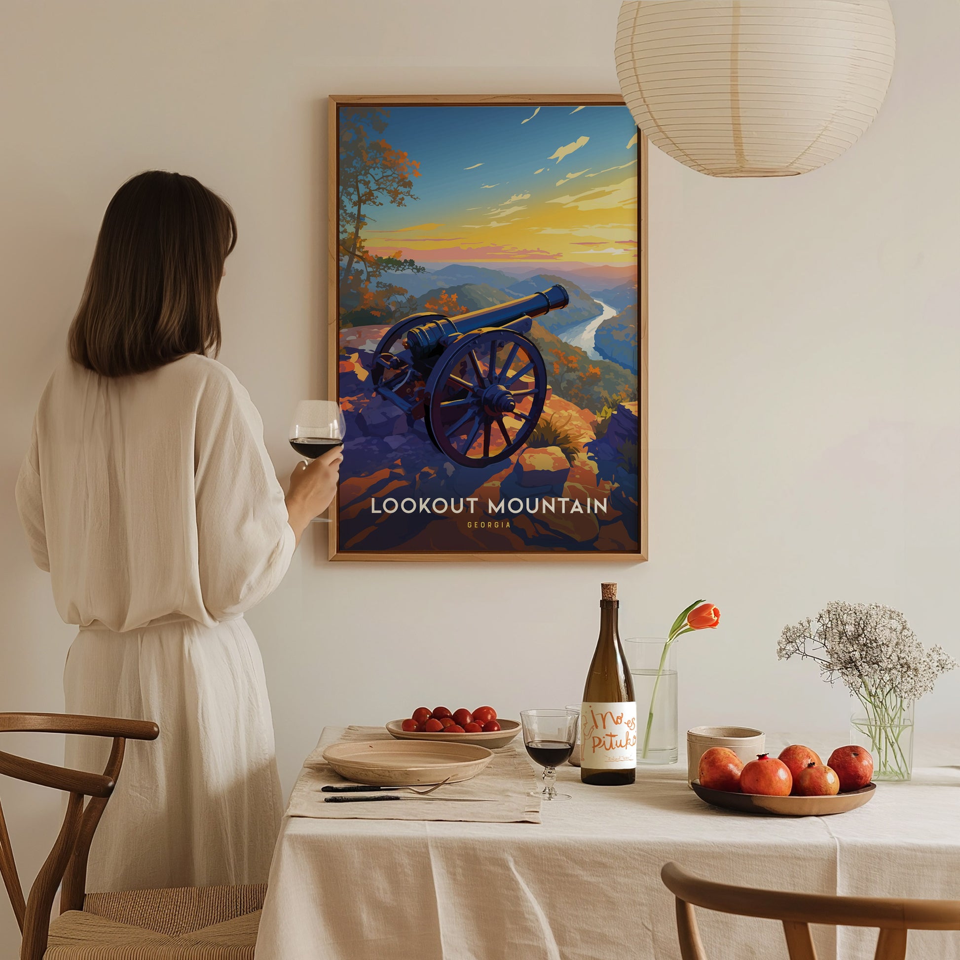 A woman in a white dress holds a glass of wine while admiring a framed Lookout Mountain poster. A dining table with fruit, flowers, and wine is in the foreground.