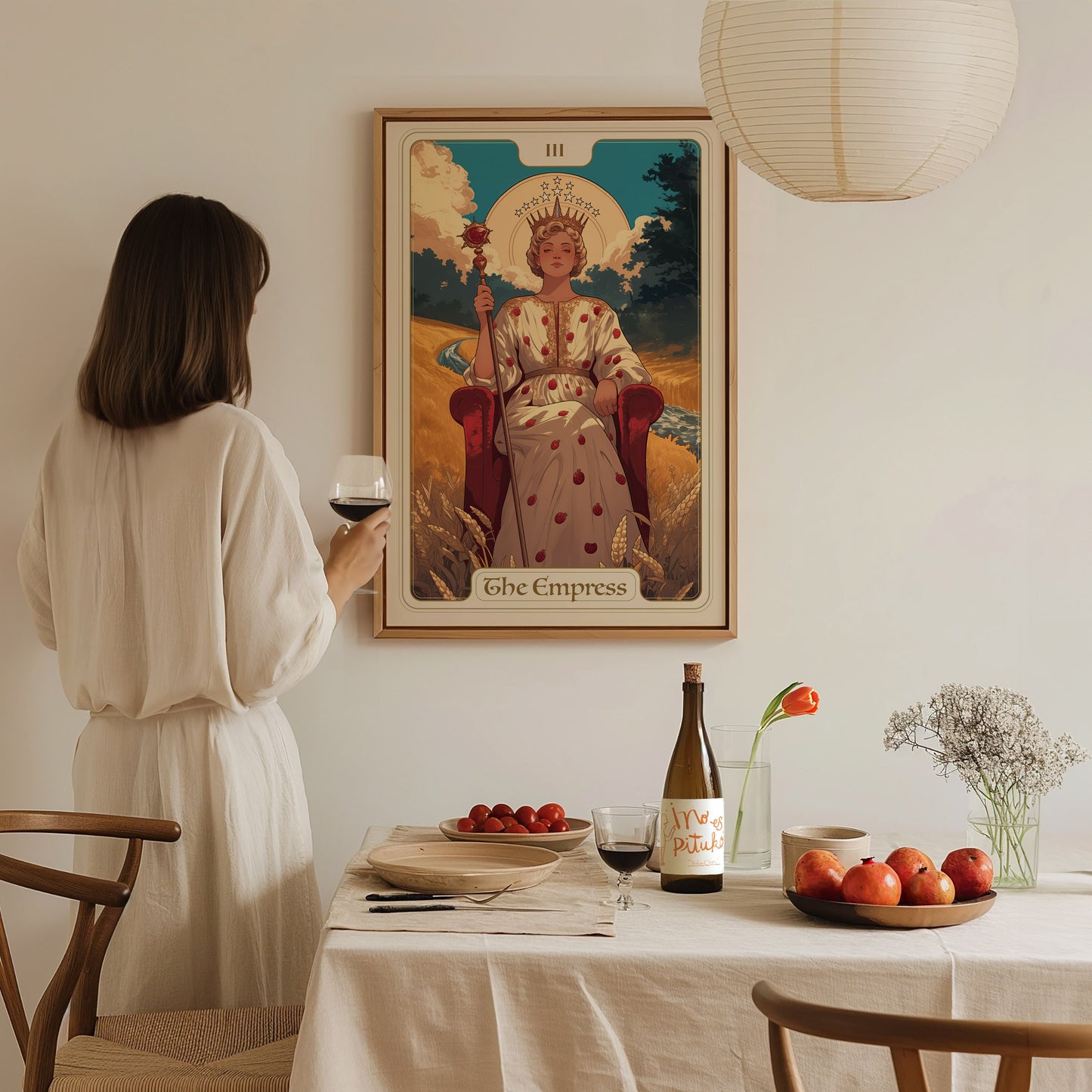 A woman in a white dress admires an Empress Tarot poster on the wall, with a dining table set with wine, fruit, and flowers in the foreground.