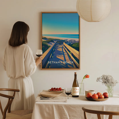 A woman in a beige dress admires a Bethany Beach poster on the wall, holding a glass of wine. A dining table with fruit and wine is in the foreground.