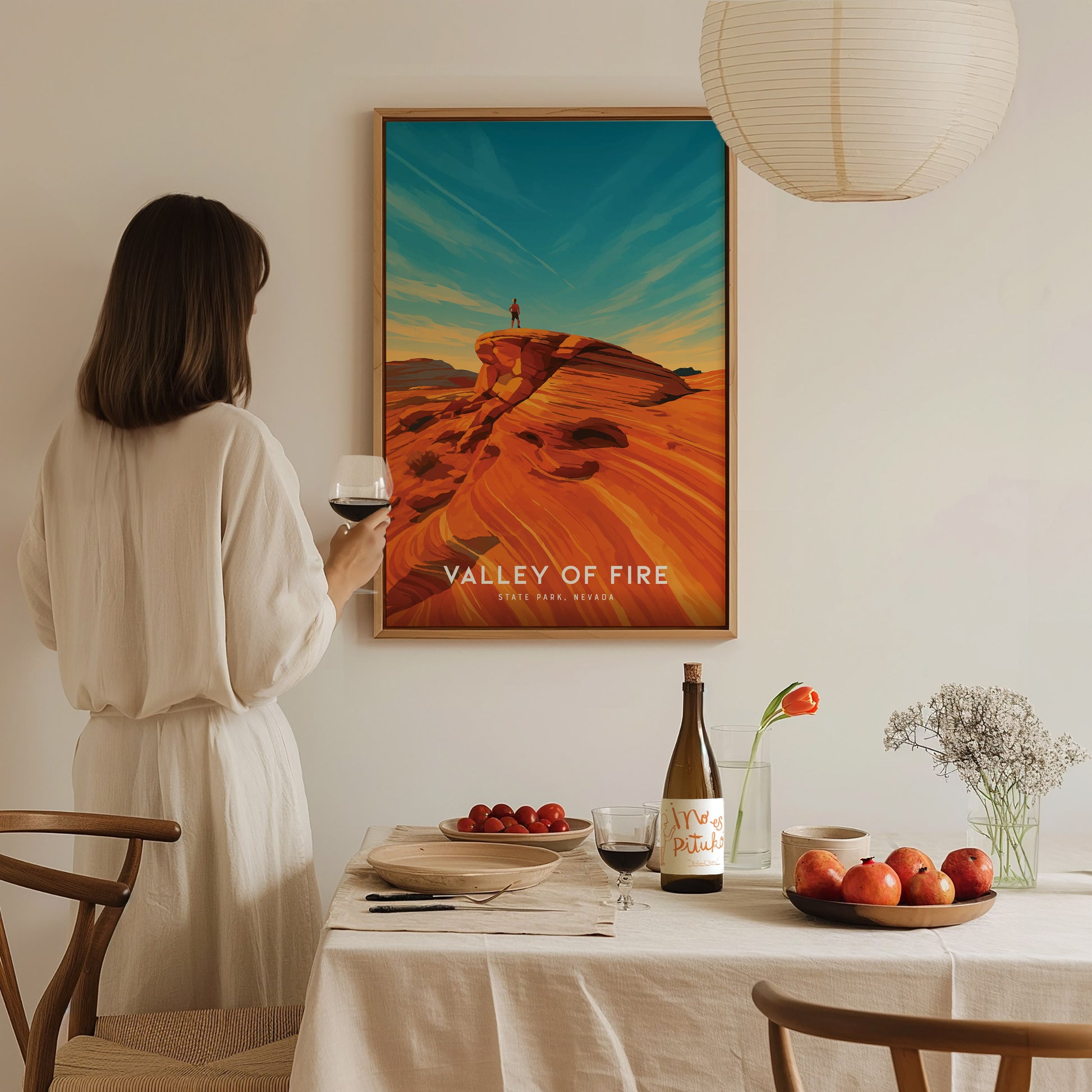 A woman in a light-colored dress stands near a table set with a bottle of wine, fruit, and a vase of flowers, gazing at a colorful poster of the Valley of Fire.