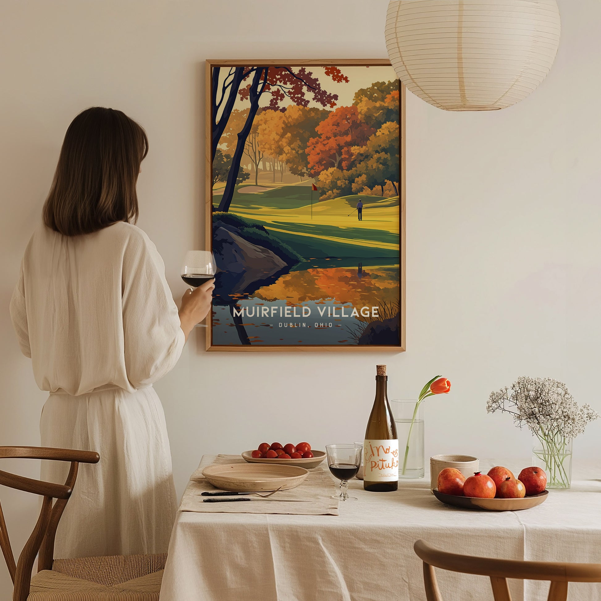 A woman in a white dress holds a glass of wine, admiring a framed Muirfield Village Golf Club poster. The dining table is set with fruit, wine, and flowers.