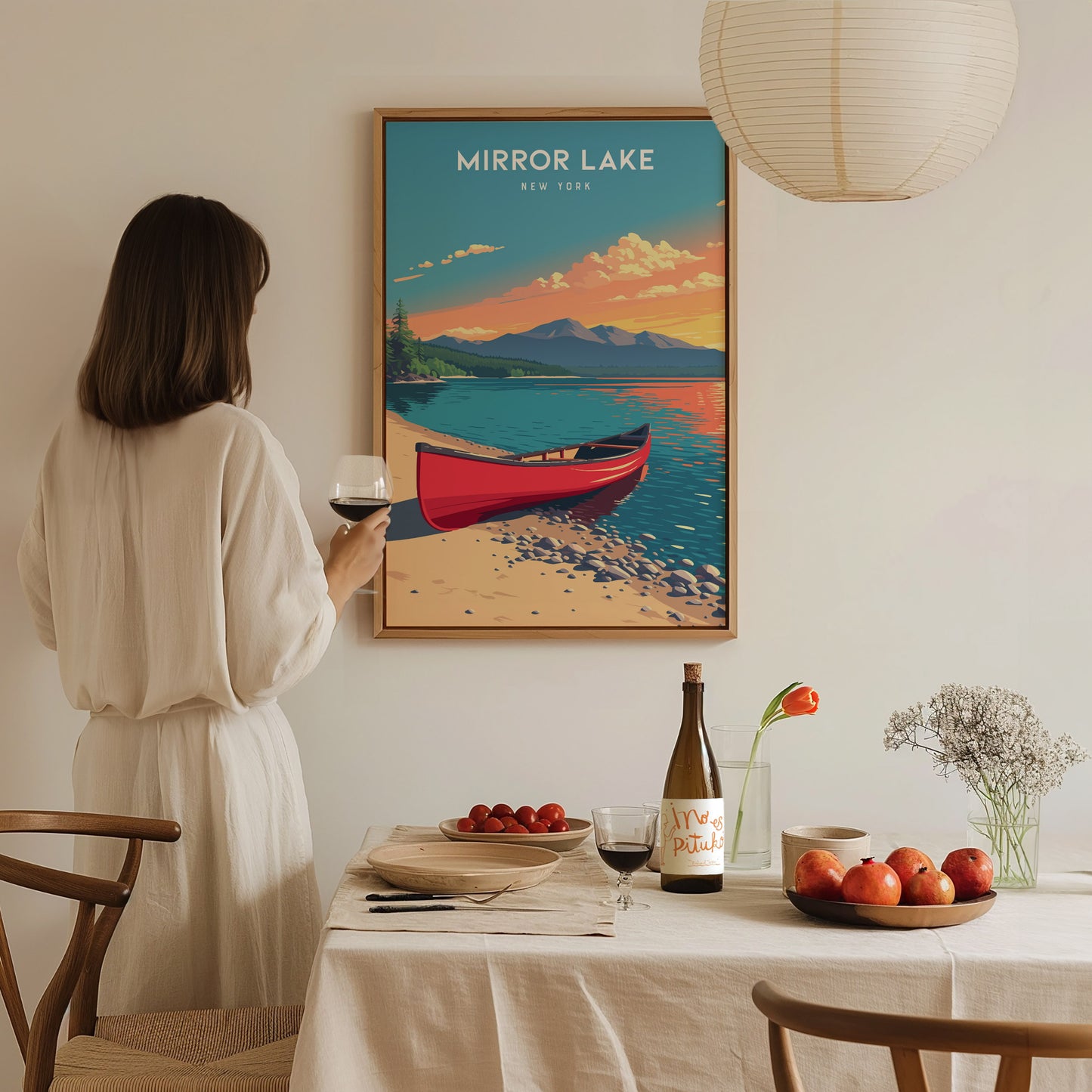 A woman in a light-colored outfit holds a glass of wine while standing near a dining table set with a bottle and fruit.