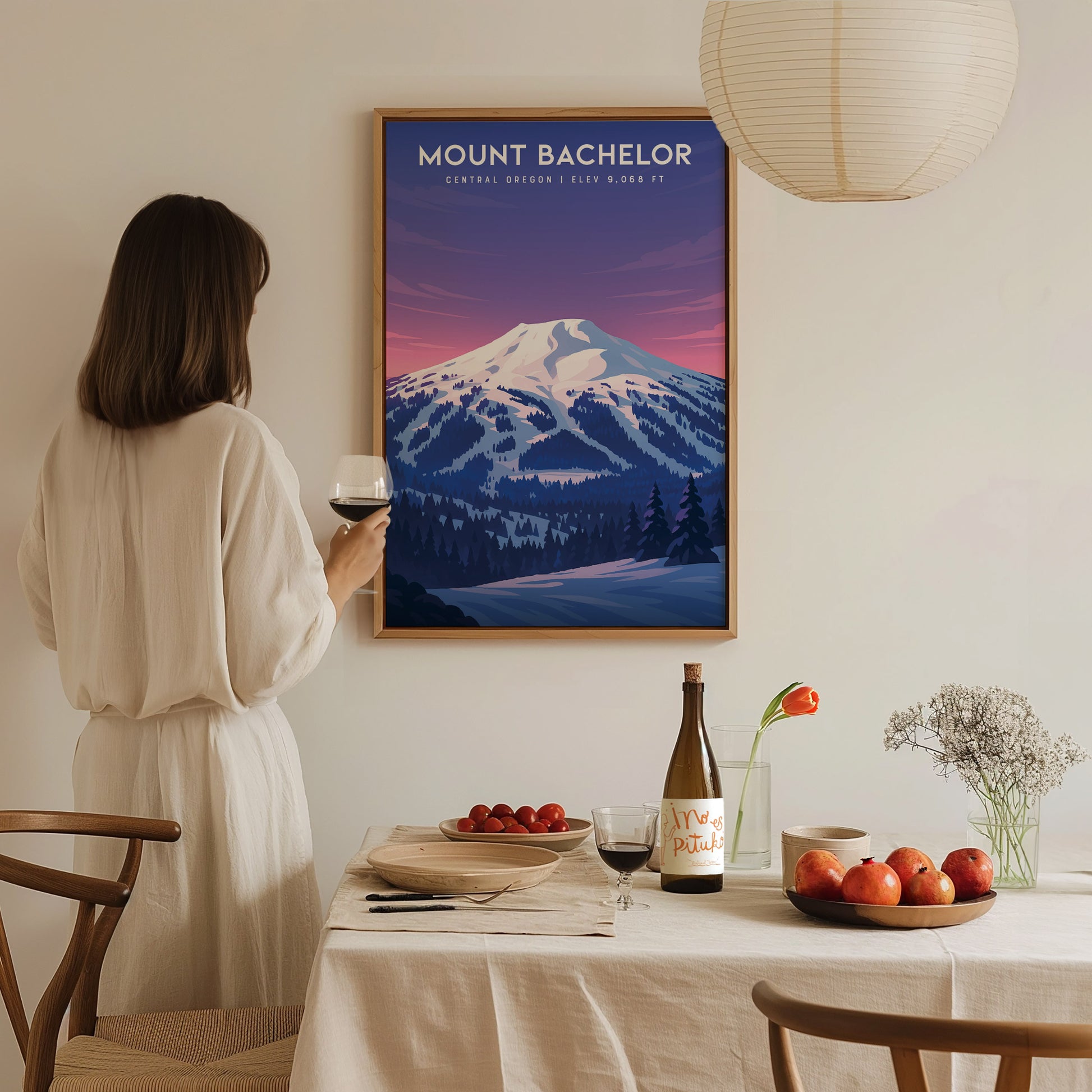 A woman in a light-colored dress stands at a dining table, holding a glass of wine, while admiring a framed art print of Mount Bachelor.