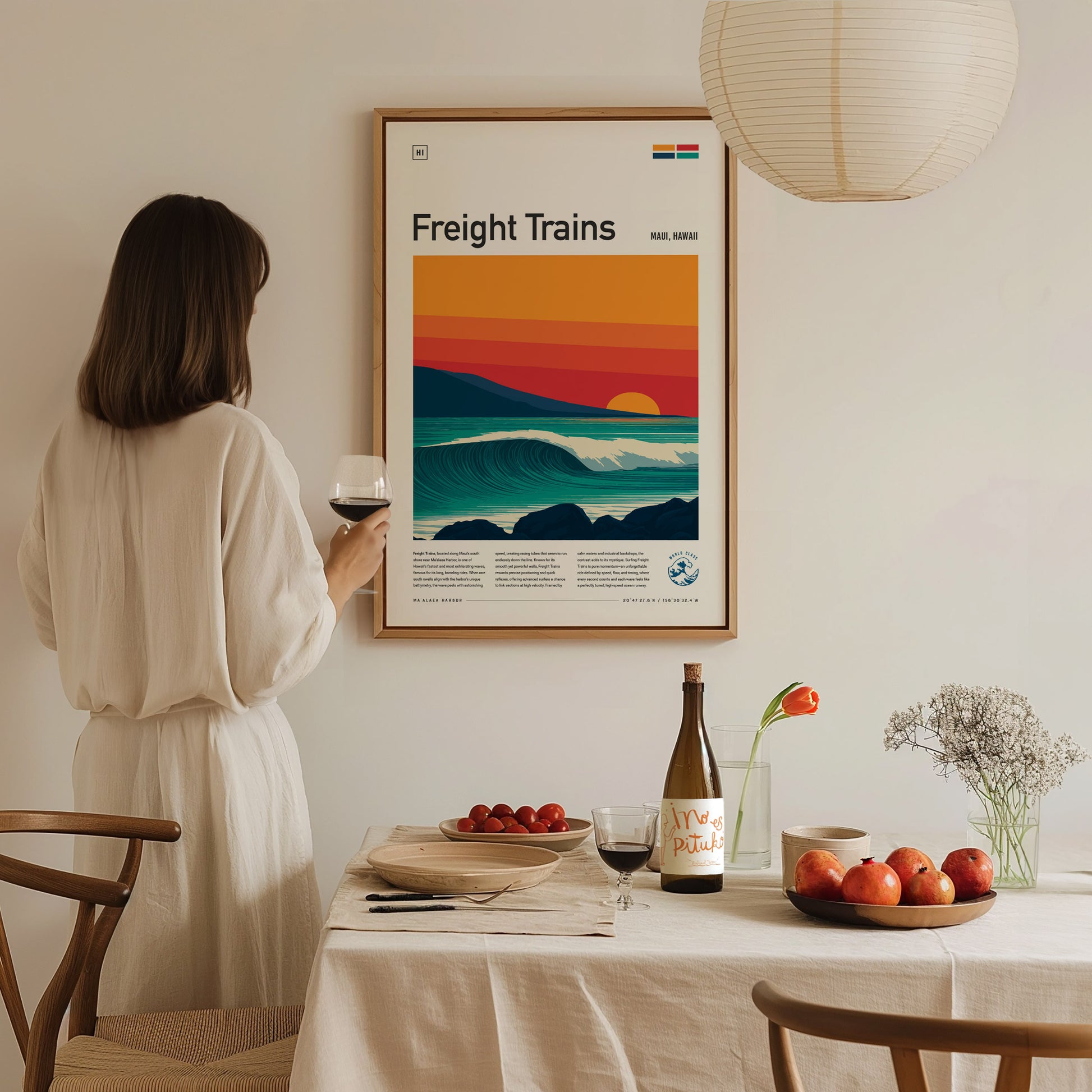 A woman in a white dress stands holding a glass of wine, gazing at a framed poster titled Freight Trains, featuring a colorful sunset over stylized waves. The scene is set in a cozy dining area with a table adorned with apples and a bottle.