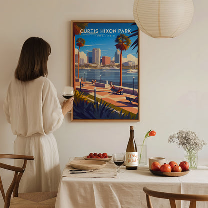 A woman in a white dress holds a glass of wine, admiring a colorful Curtis Hixon Park poster. A table with fruit, wine, and flowers is in the foreground.