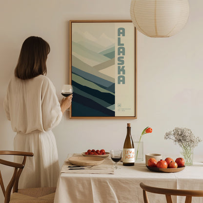 A woman in a beige dress holds a glass of wine, admiring a framed Alaska poster with abstract mountain design. A dining table with fruits and a wine bottle is.