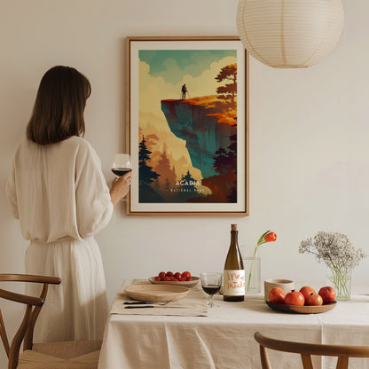 A woman in a beige dress holds a glass of wine, gazing at a framed Acadia National Park poster. The minimalist artwork features dramatic cliffs and trees.
