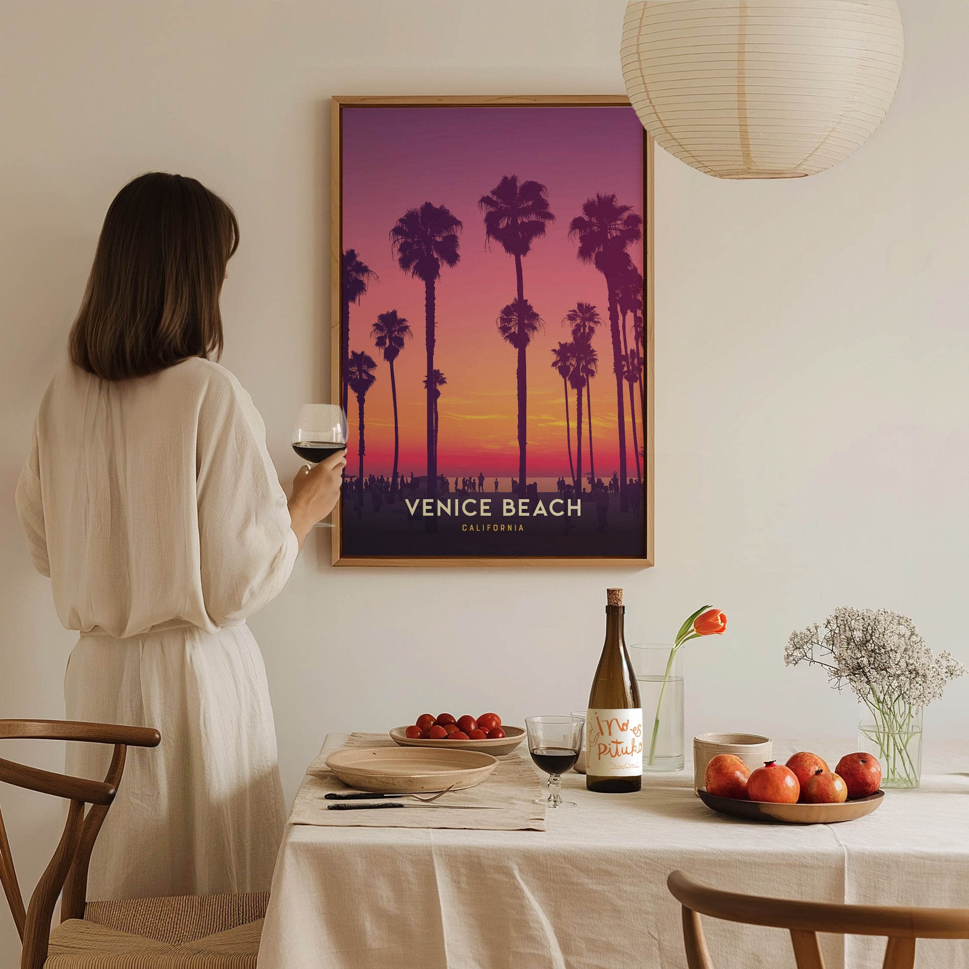 A woman in a white dress admires a framed Venice Beach sunset poster with palm trees. A table with wine, fruit, and flowers is set in the foreground.