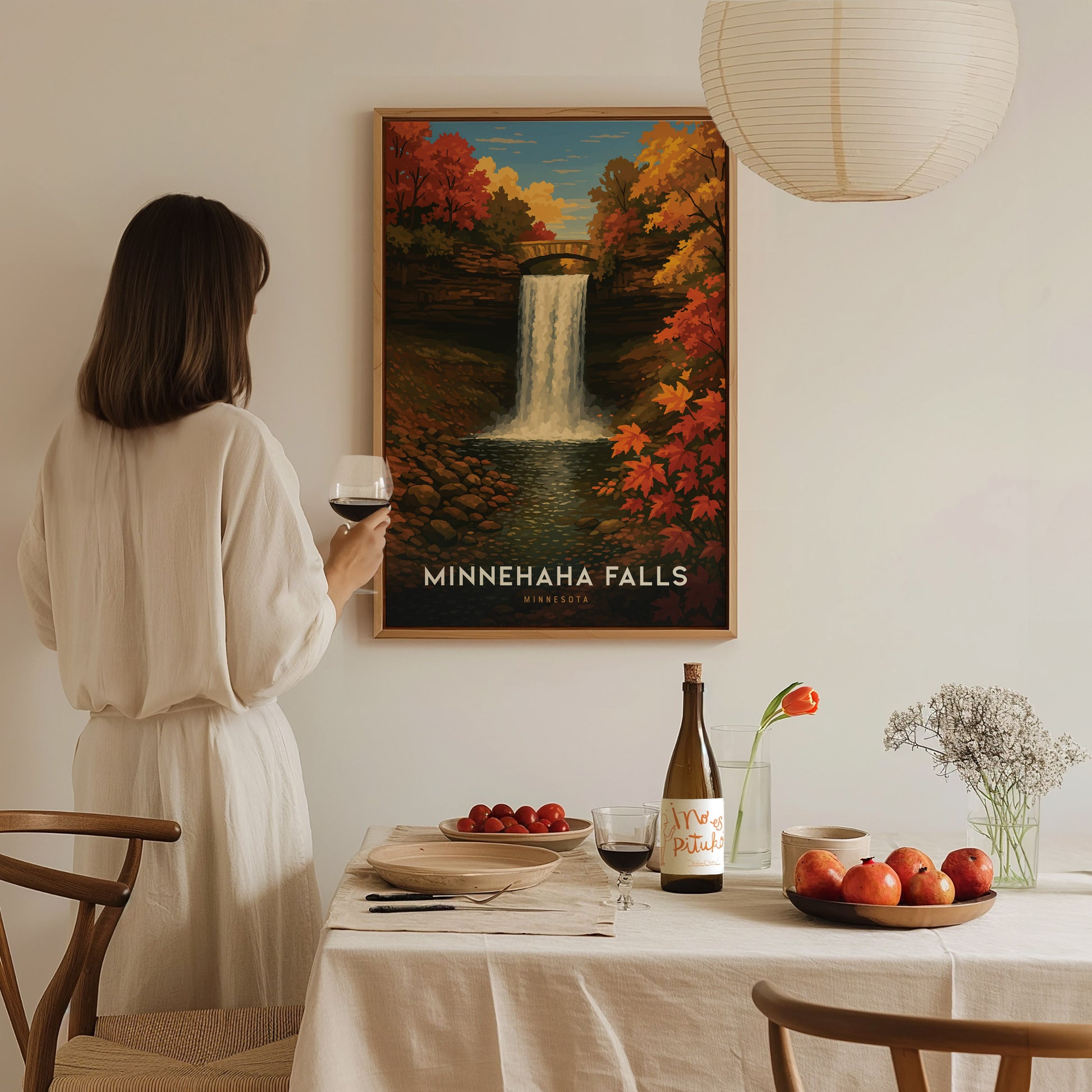 A person in a white dress holds a glass of wine while looking at a Minnehaha Falls poster. A table with fruit, flowers, and wine is in the foreground.