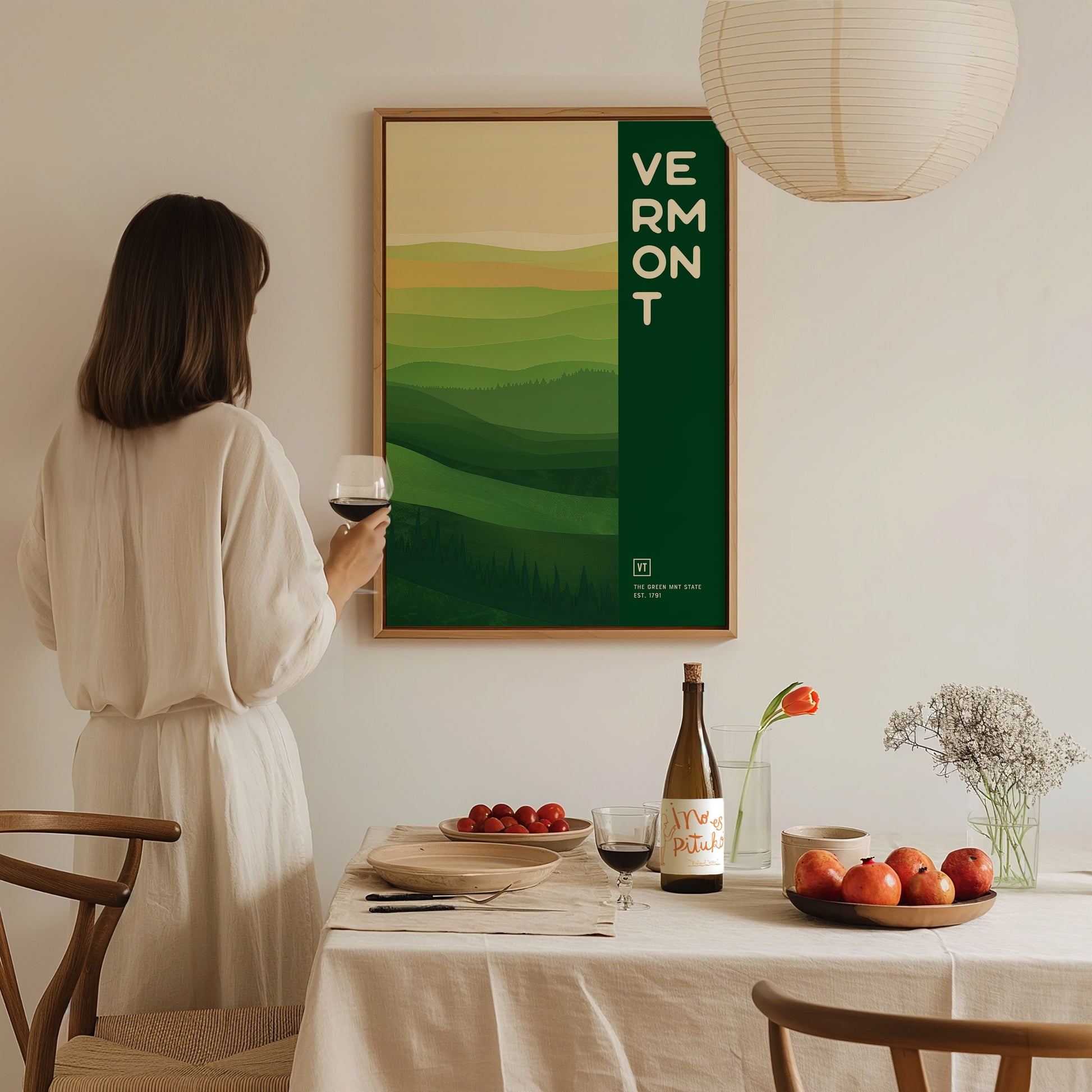 A woman in a white dress holding a wine glass observes a Vermont poster with green hills. A table with fruits, a wine bottle, and flowers is nearby.