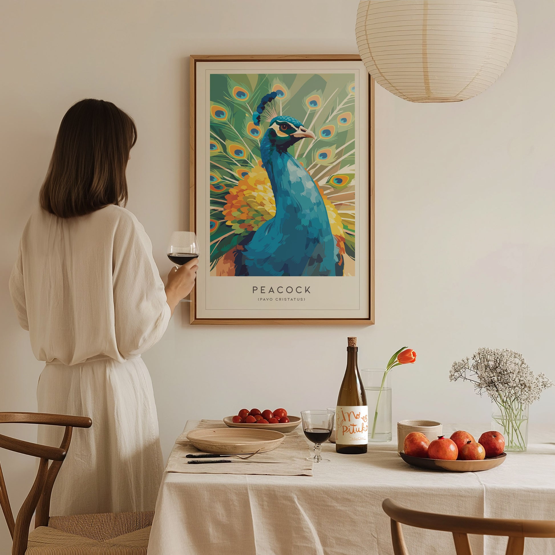 A woman in a white dress stands near a dining table, admiring a colorful peacock art print on the wall. The table is set with a bottle of wine, a plate of apples, and a small vase of flowers, creating a warm and inviting atmosphere.