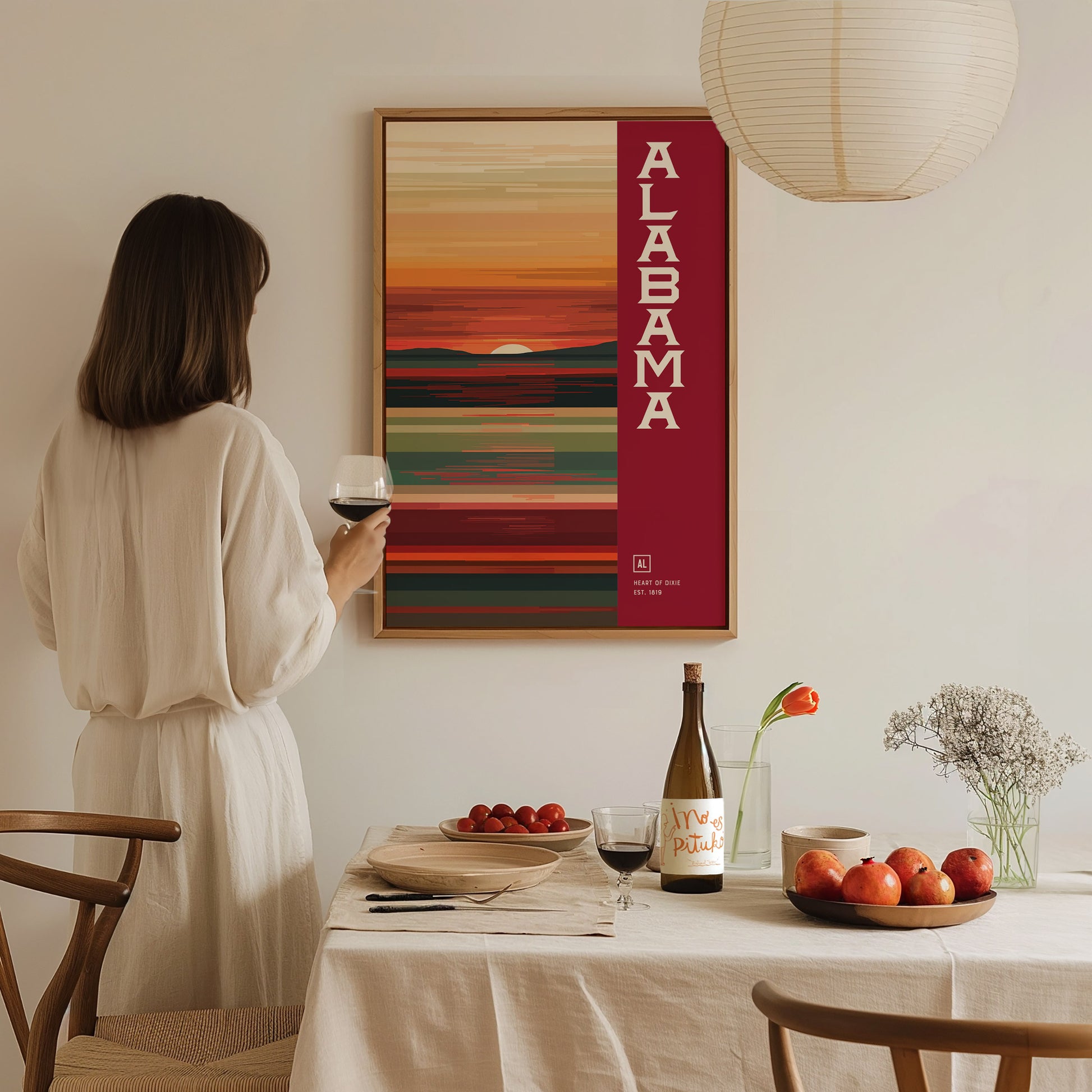 A woman in a white dress holds a glass of wine, admiring a framed Alabama abstract poster on the wall. A dining table with fruit and a wine bottle is.