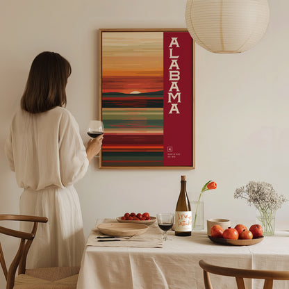 A woman in a white dress holds a glass of wine, admiring a framed Alabama abstract poster on the wall. A dining table with fruit and a wine bottle is.