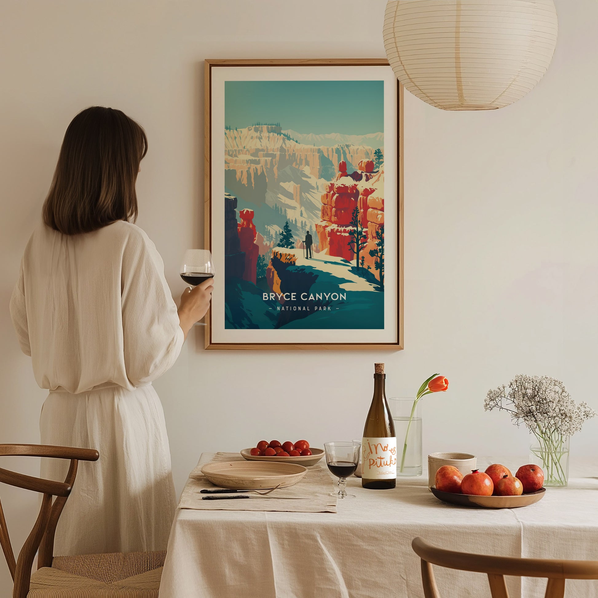 A woman in a white dress holds a glass of wine, gazing at a framed Bryce Canyon National Park print. A table with fruit and wine is in the foreground.