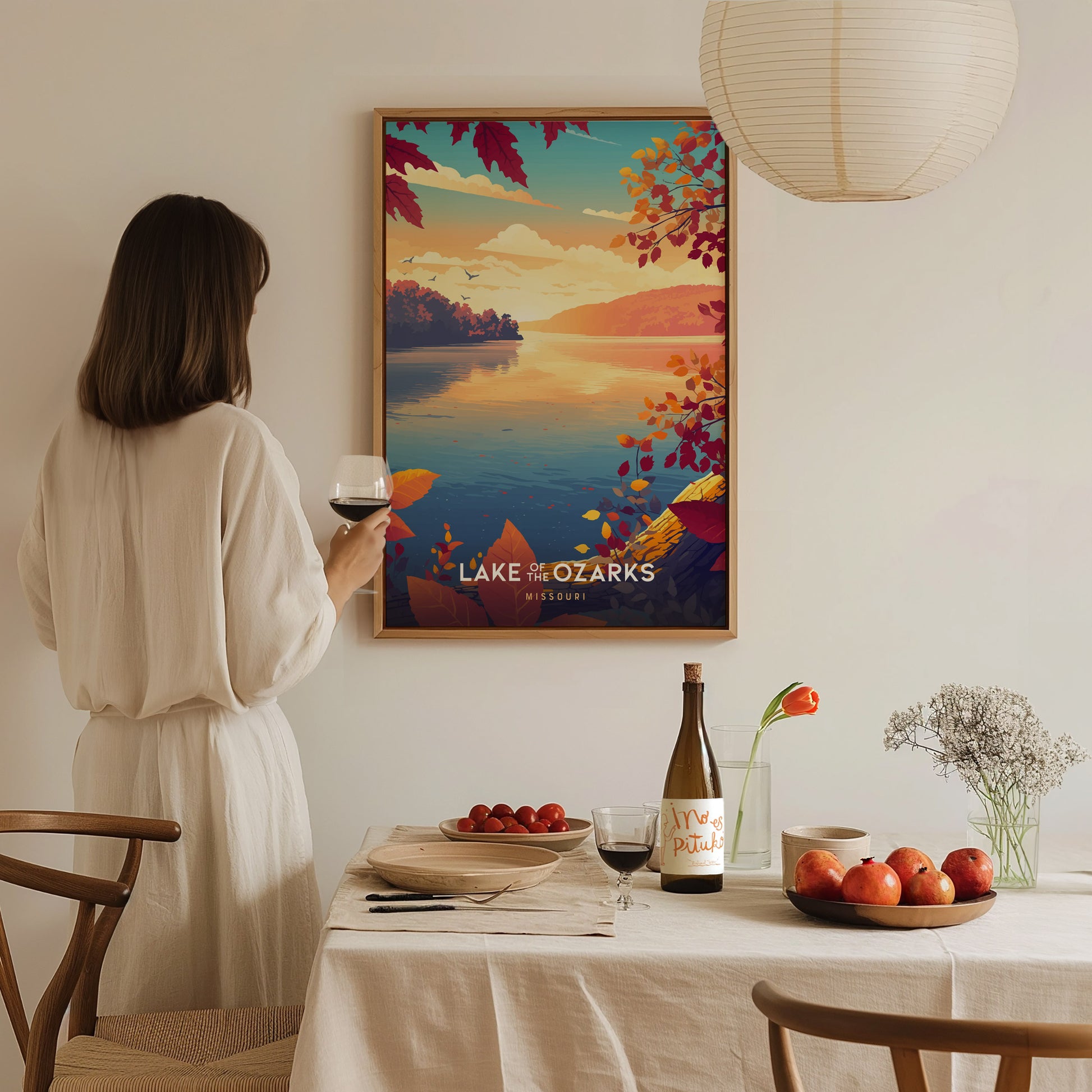 A woman in a light-colored dress stands in a softly lit room, holding a glass of wine while gazing at a colorful poster of Lake of the Ozarks.