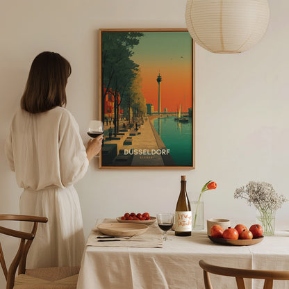 A woman in a white dress stands at a dining table, holding a glass of wine, gazing at a framed poster of Düsseldorf featuring a scenic canal view with trees and a tower in the background.