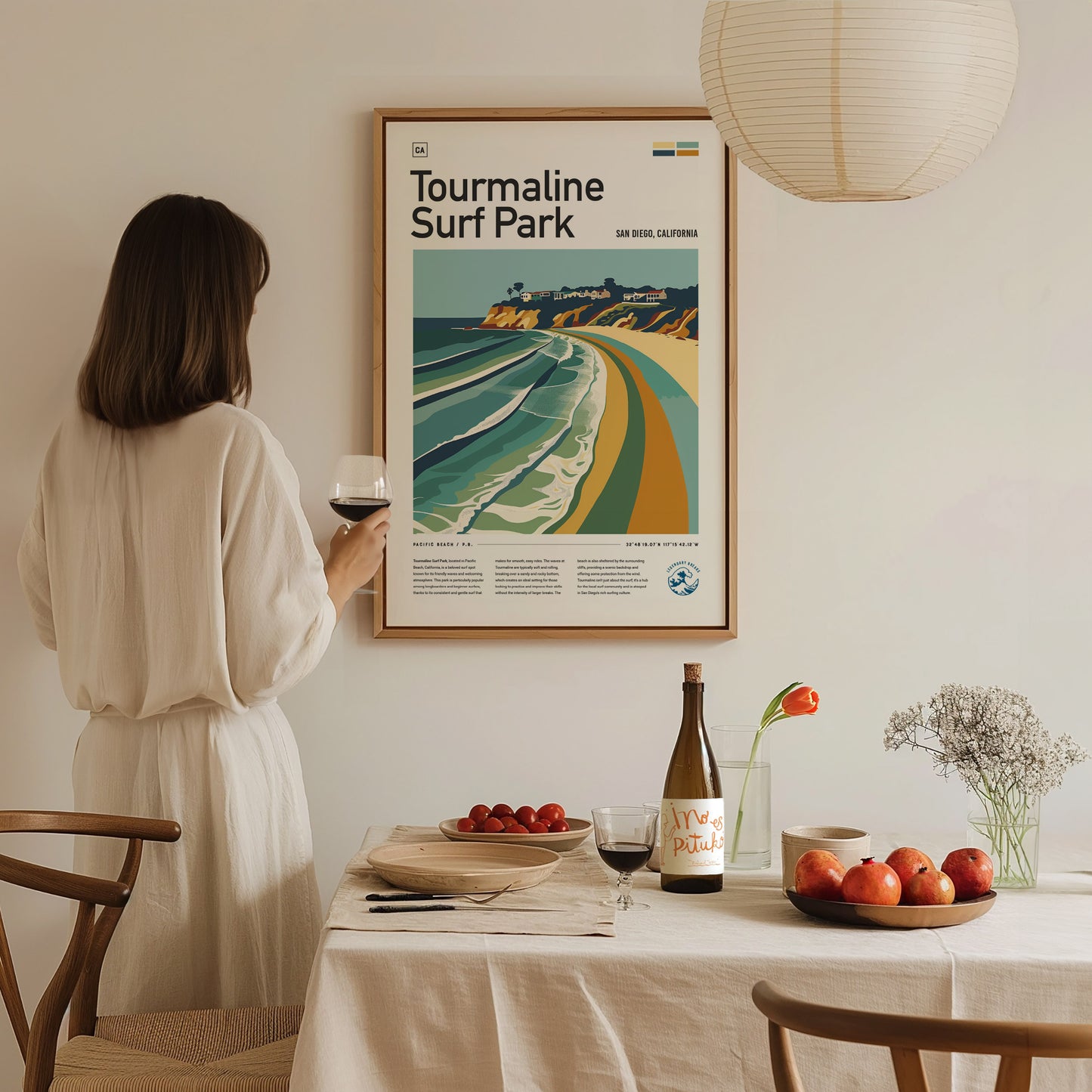 A woman in a white dress admires a framed Tourmaline Surf Park poster. A dining table with wine, fruits, and flowers is set beneath a hanging lamp.