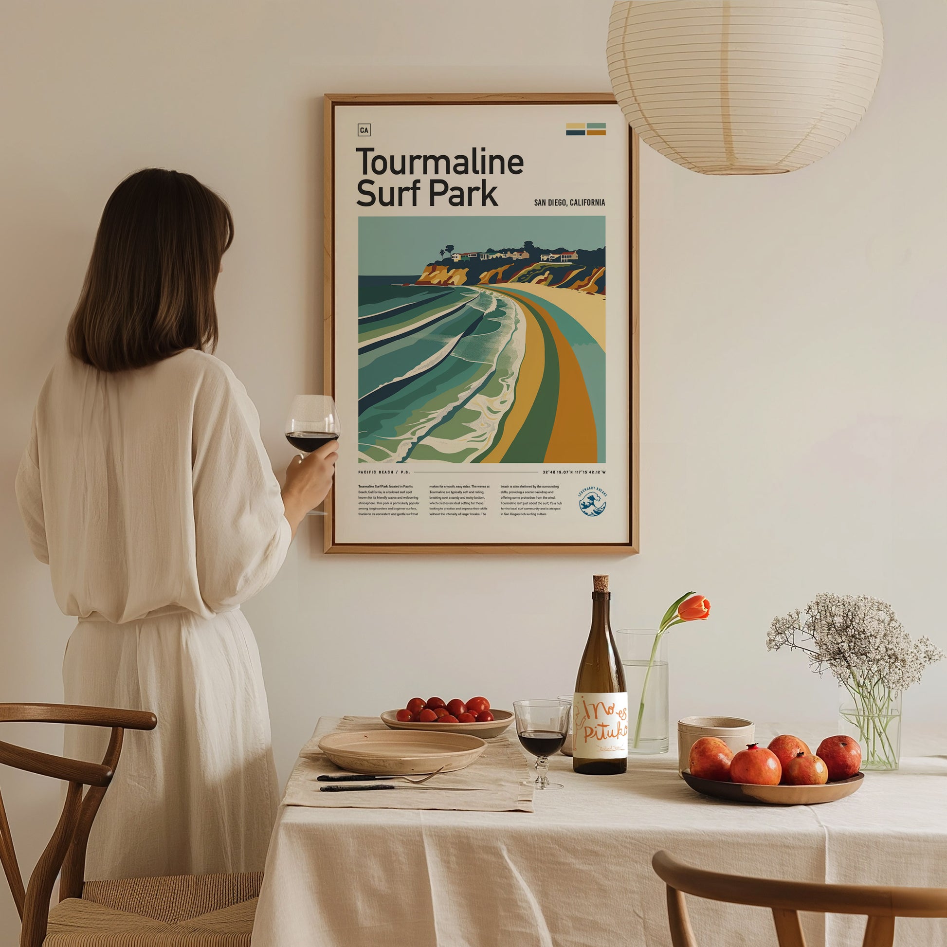 A woman in a white dress admires a framed Tourmaline Surf Park poster. A dining table with wine, fruits, and flowers is set beneath a hanging lamp.