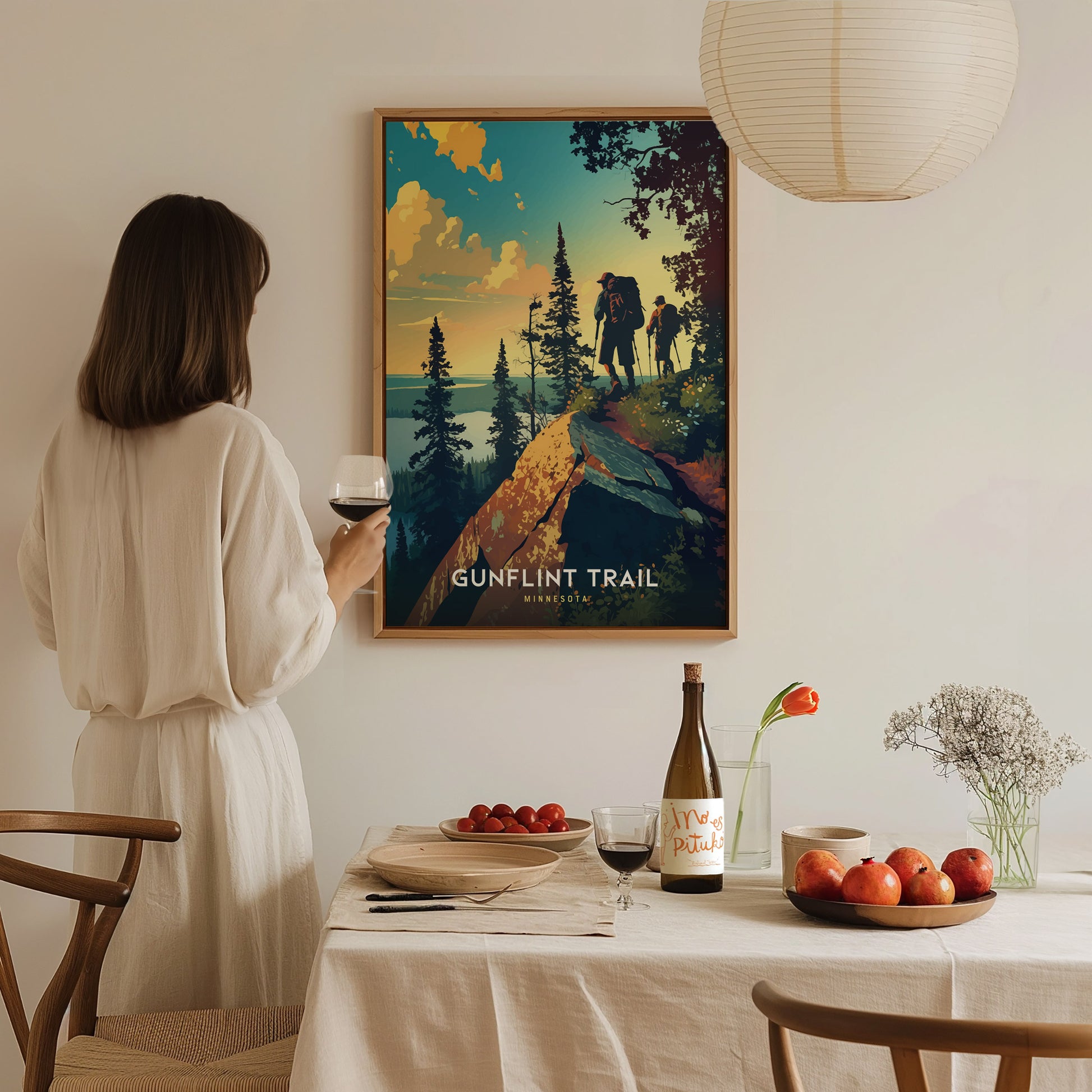 A woman in a white dress holds a glass of wine, admiring a Gunflint Trail poster on the wall. A table with fruit, flowers, and wine is in the foreground.
