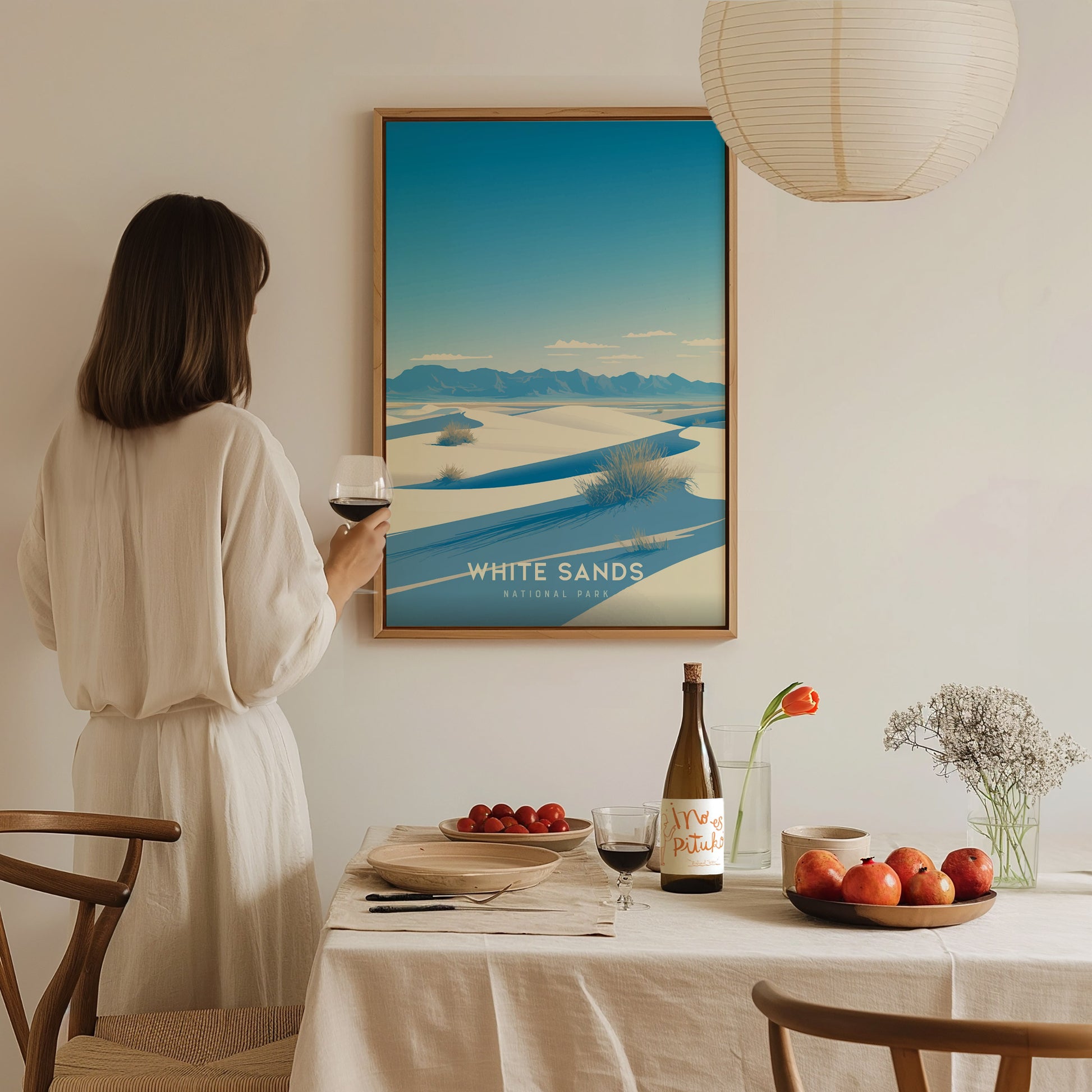 A woman in a white dress holds a glass of wine, admiring a framed White Sands National Park poster. A table with fruit, wine, and flowers is in the foreground.