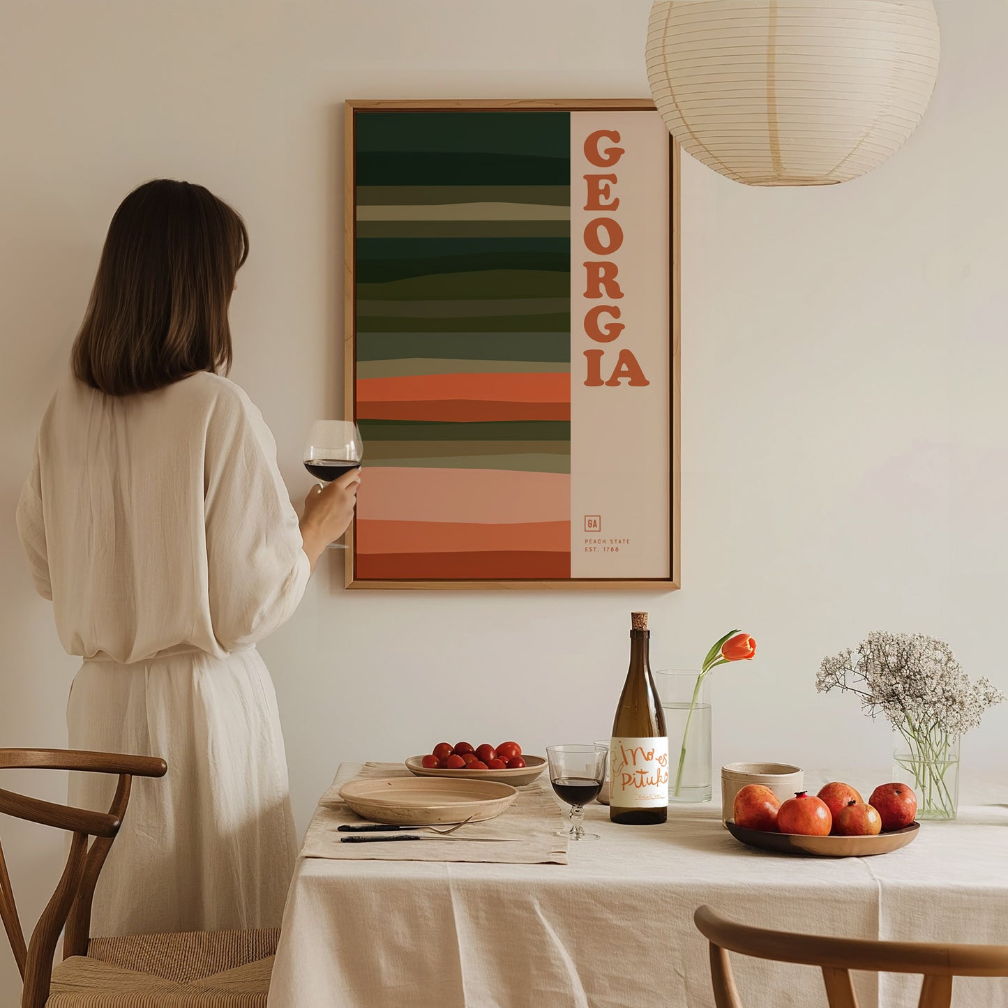 A woman in a white dress holds a wine glass while gazing at a colorful Georgia poster on the wall. A table is set with a bottle, fruit, and flowers.