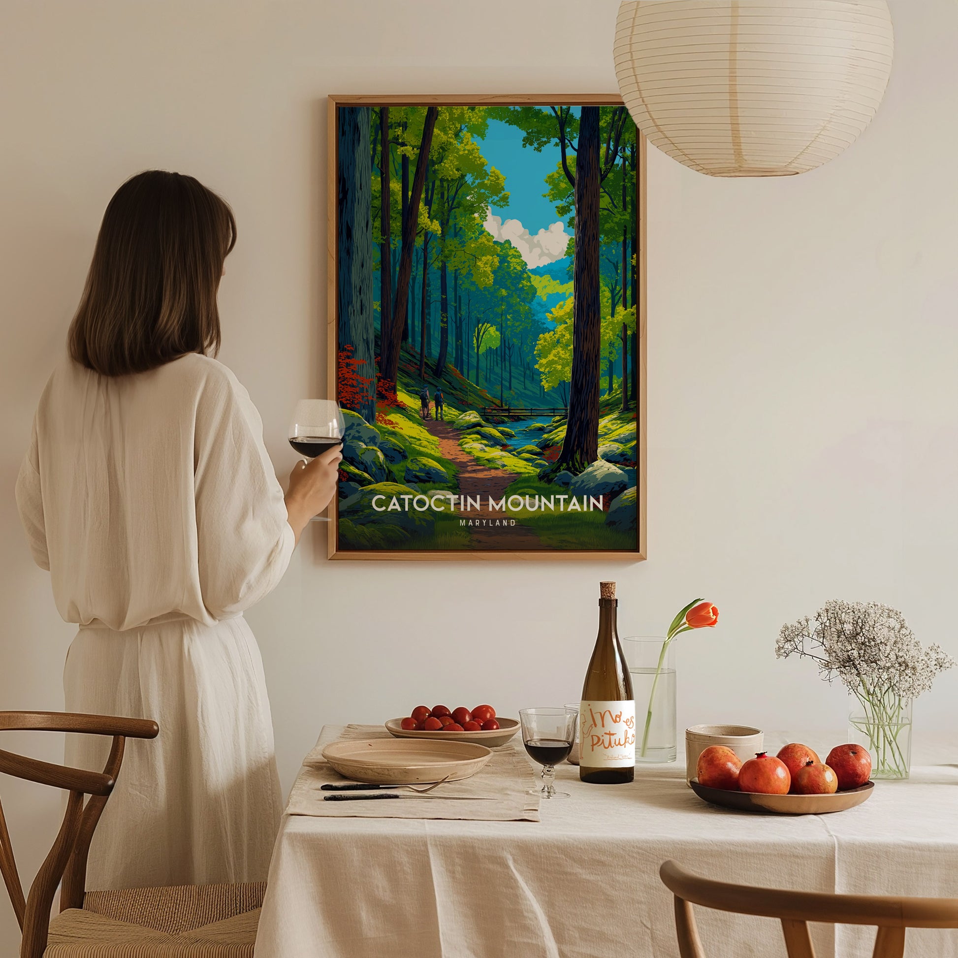 A woman in a white dress stands at a dining table, holding a glass of red wine and gazing at a vibrant poster of Catoctin Mountain, depicting a lush forest path.