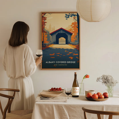 A woman stands in a cozy dining area, holding a glass of wine and admiring a framed poster of the Albany Covered Bridge in New Hampshire.