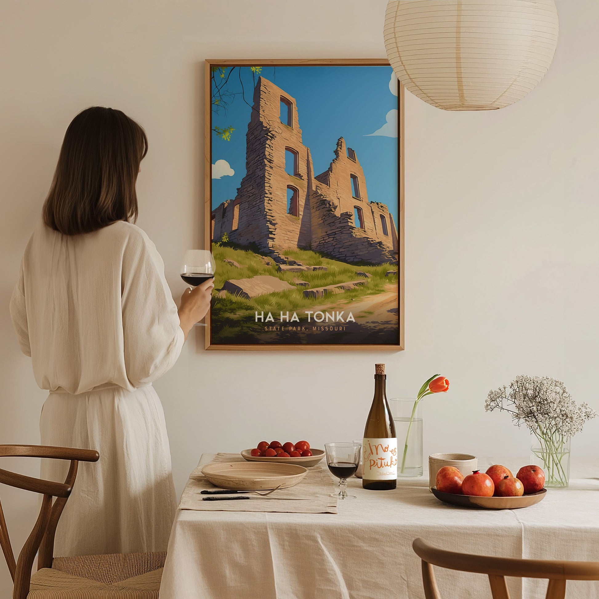 A woman in a light-colored dress stands sipping wine beside a dining table adorned with fruits and a bottle, gazing at a framed art print of the Ha Ha Tonka State Park castle ruins. The scene is warmly lit, creating a cozy atmosphere.