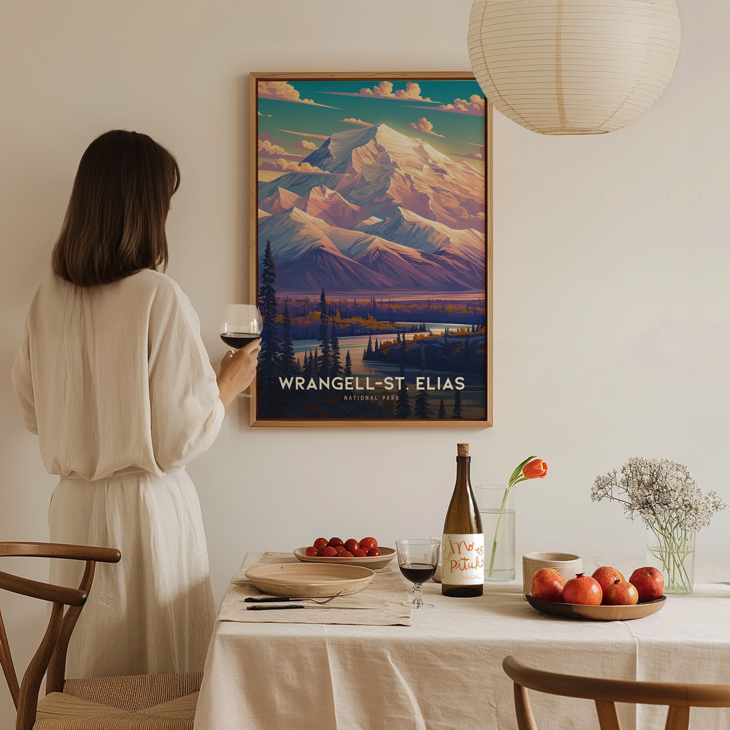 A person in a white dress holds a glass of wine while admiring a Wrangell-St. Elias National Park poster. A dining table with fruit and wine is in the foreground.