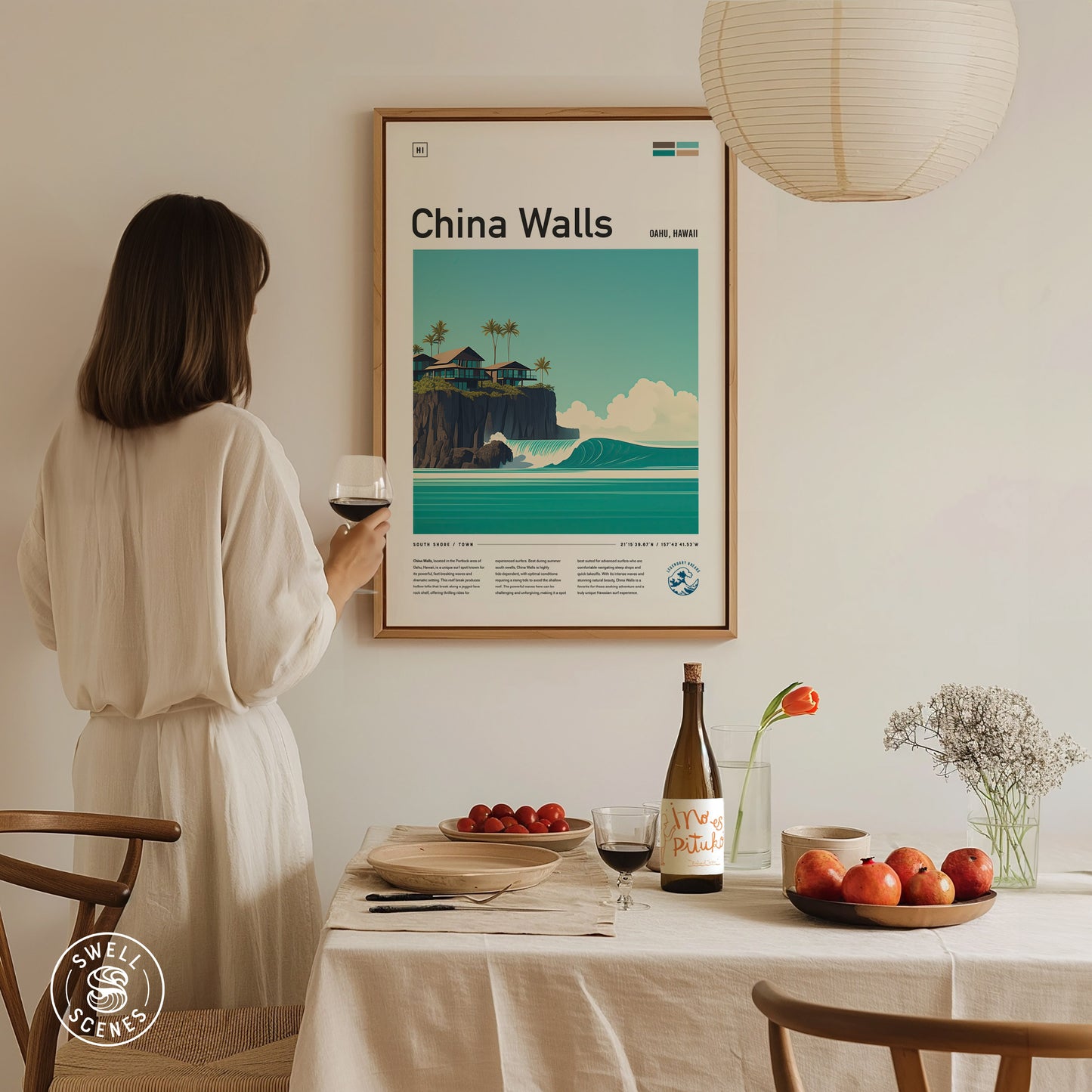 A woman in a white dress holds a wine glass, admiring a framed China Walls poster. A table with apples, flowers, and wine is set beneath a hanging lamp.