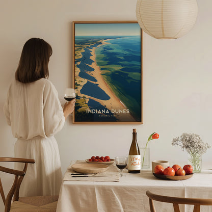 A woman in a white dress holds a glass of wine, admiring a framed Indiana Dunes National Park poster. A table in the foreground features fruit, a bottle, and flowers.