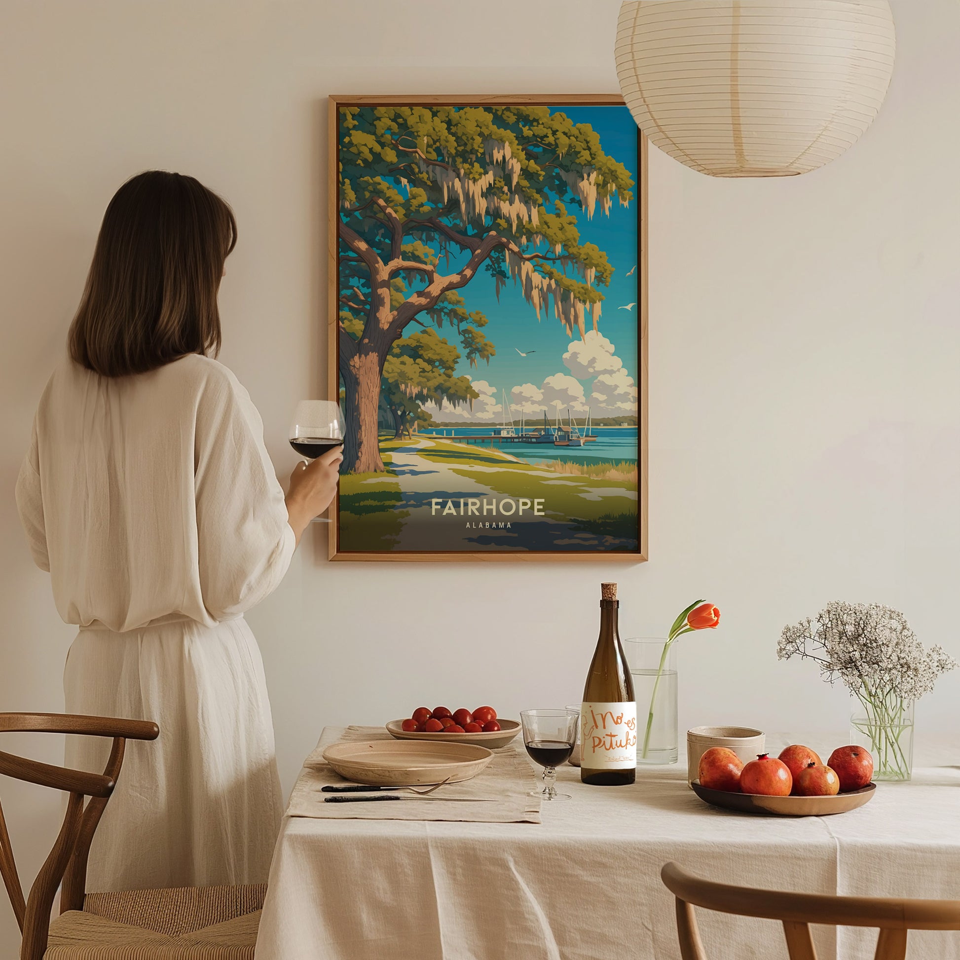 A woman in a white dress holds a glass of wine, admiring a framed Fairhope, Alabama poster. A table with fruit, wine, and flowers is in the foreground.