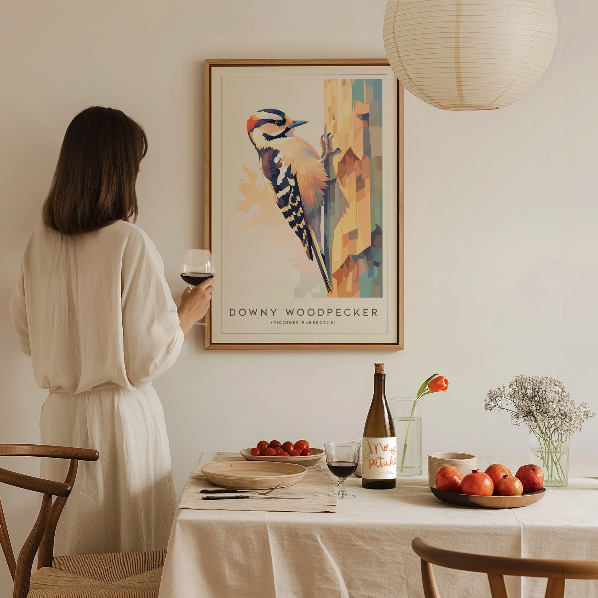 A person stands in a softly lit room, holding a glass of wine and admiring an art print of a Downy Woodpecker, which is displayed prominently on the wall.