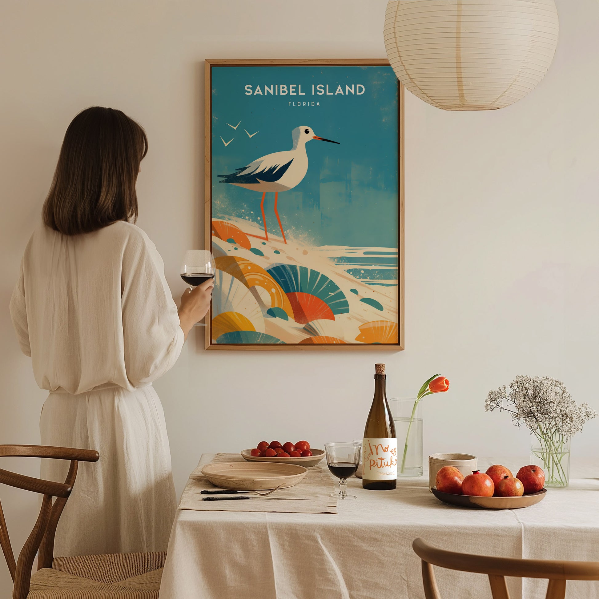 A woman in a white dress holds a glass of red wine while looking at a Sanibel Island poster featuring a sandpiper and seashells.