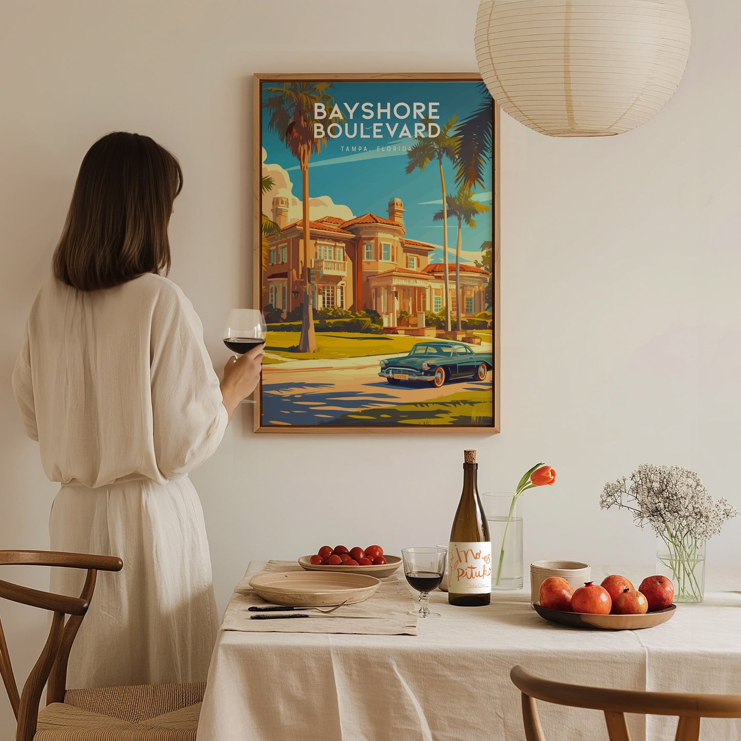 A woman in a white dress holds a glass of wine, gazing at a colorful Bayshore Boulevard poster. A dining table with fruit and wine is in the foreground.