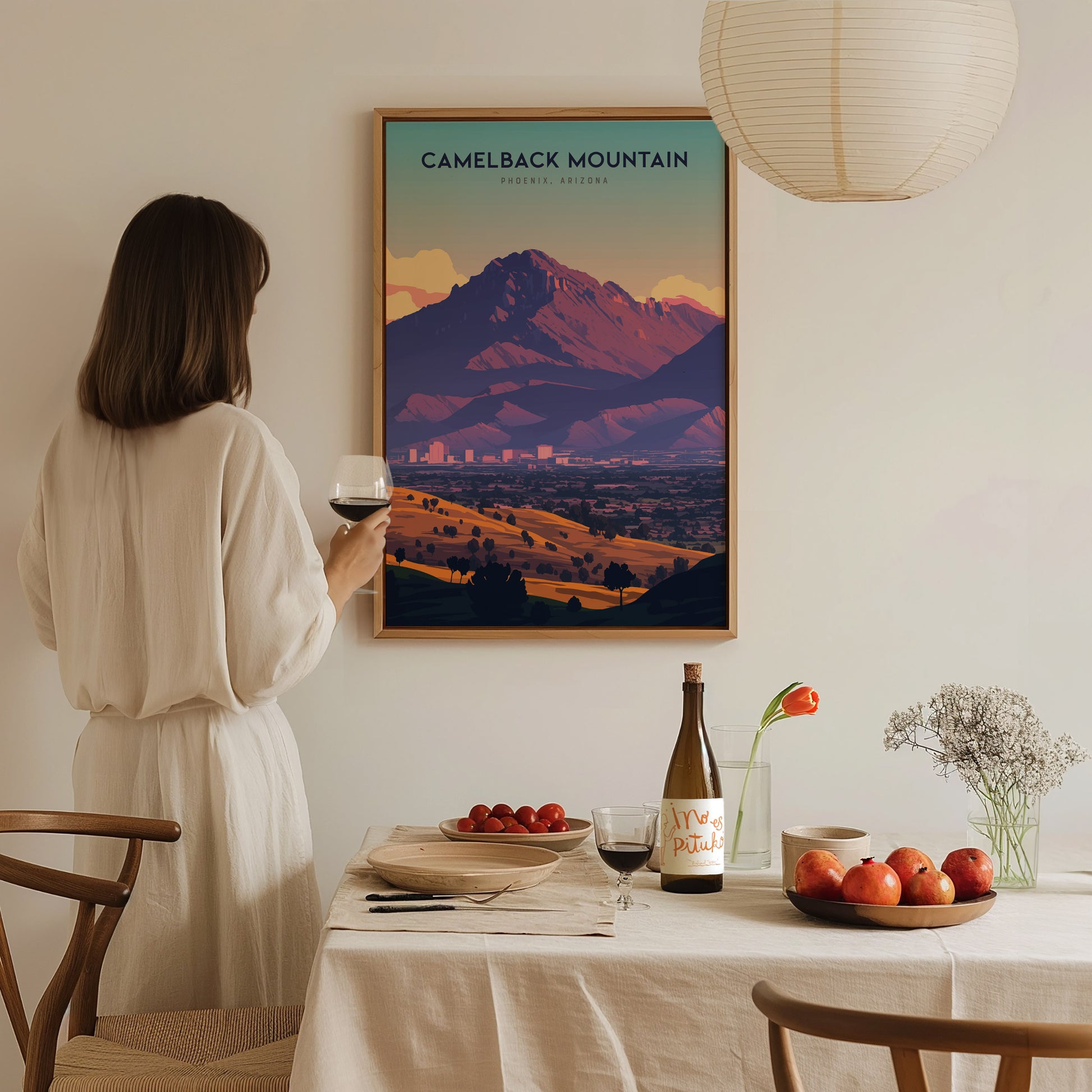 A woman in a light dress holds a glass of wine while admiring a framed Camelback Mountain poster. A table with fruit, wine, and flowers is in the foreground.