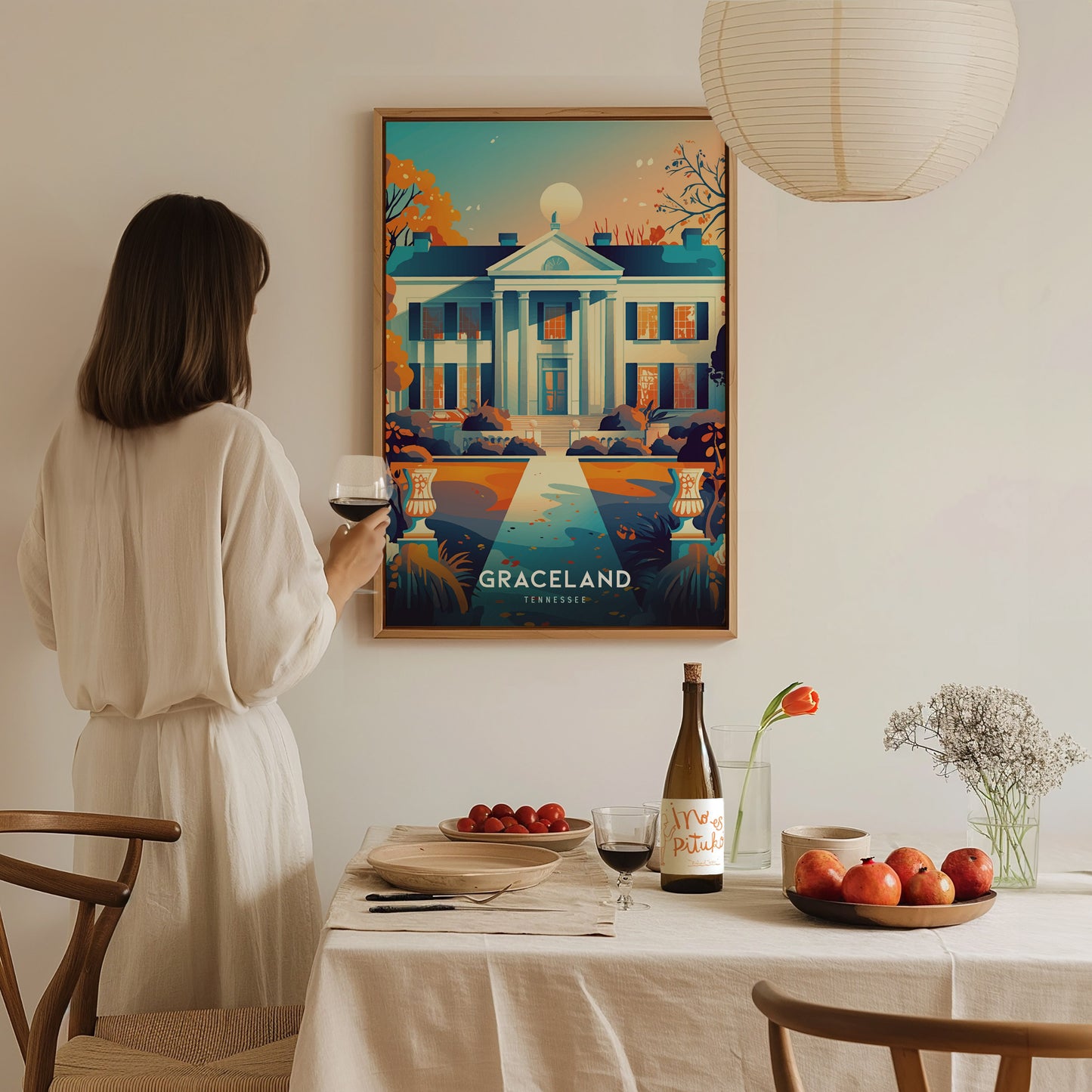 A woman in a white dress holds a glass of wine, admiring a colorful Graceland poster on the wall. A dining table below is set with fruit, flowers, and wine.