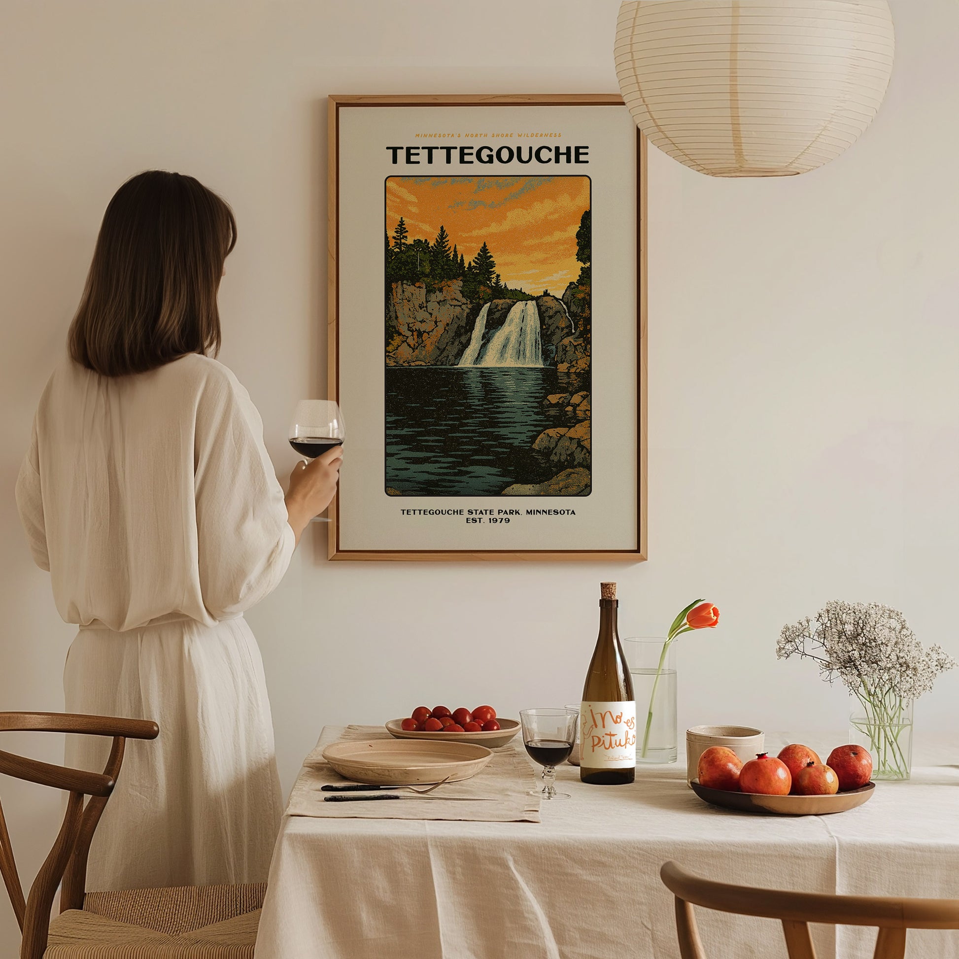 A woman in a white dress holds a glass of wine, admiring a Tettegouche State Park poster. The scene includes a set dining table with fruit and wine.