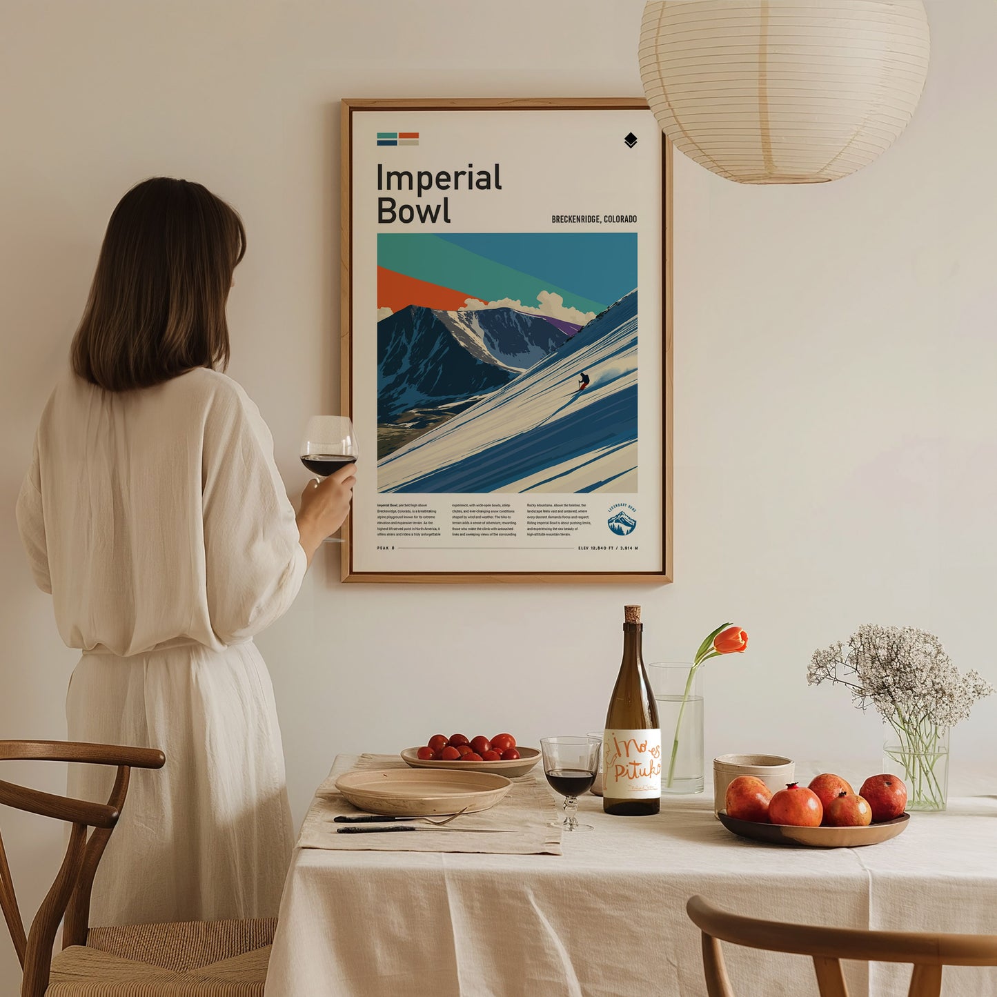 A woman in a white dress stands holding a glass of wine while gazing at a framed Imperial Bowl ski poster on the wall. The dining table in front of her is set with plates, a bottle of wine, and a bowl of apples, creating a cozy atmosphere.