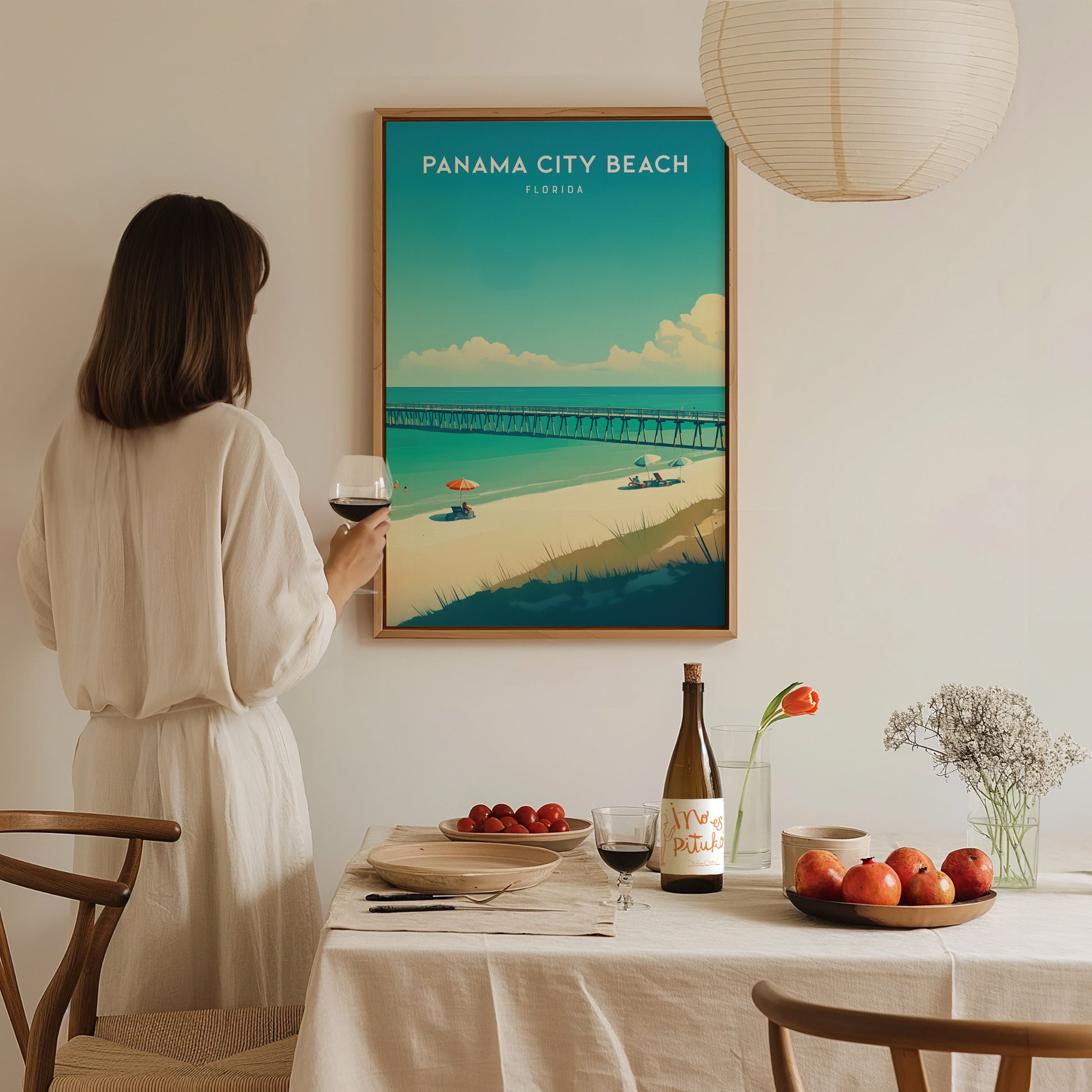 A woman in a white dress admires a Panama City Beach poster, with a dining table set with fruit, wine, and flowers in the foreground.