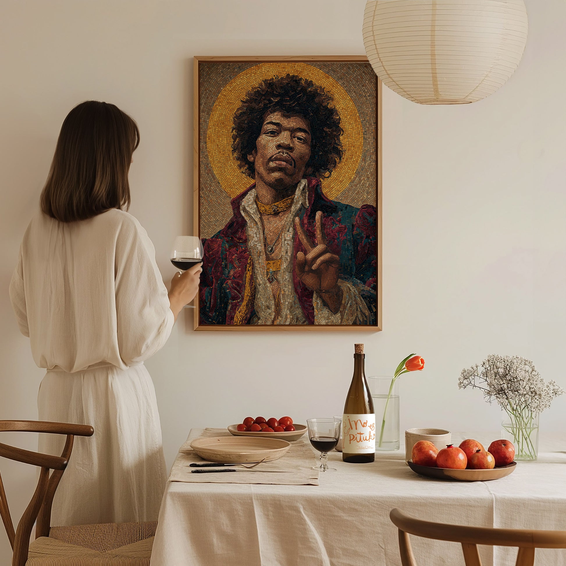 A woman in a white outfit stands at a dining table, holding a glass of wine while gazing at a framed mosaic-style portrait of a man making a peace sign. The warm, minimalist setting features a wooden table with apples and a bottle of wine.