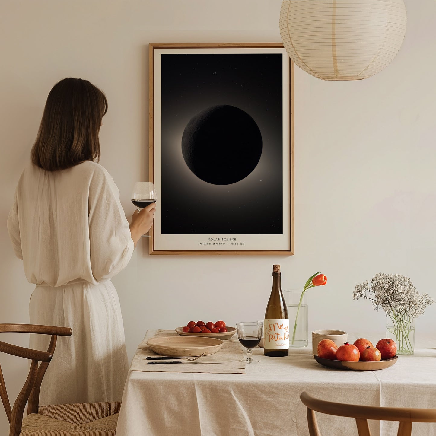 A woman in a white outfit holds a glass of wine while standing beside a dining table set with fruit and a bottle. On the wall, theres a framed poster depicting a solar eclipse, creating a serene, elegant atmosphere.