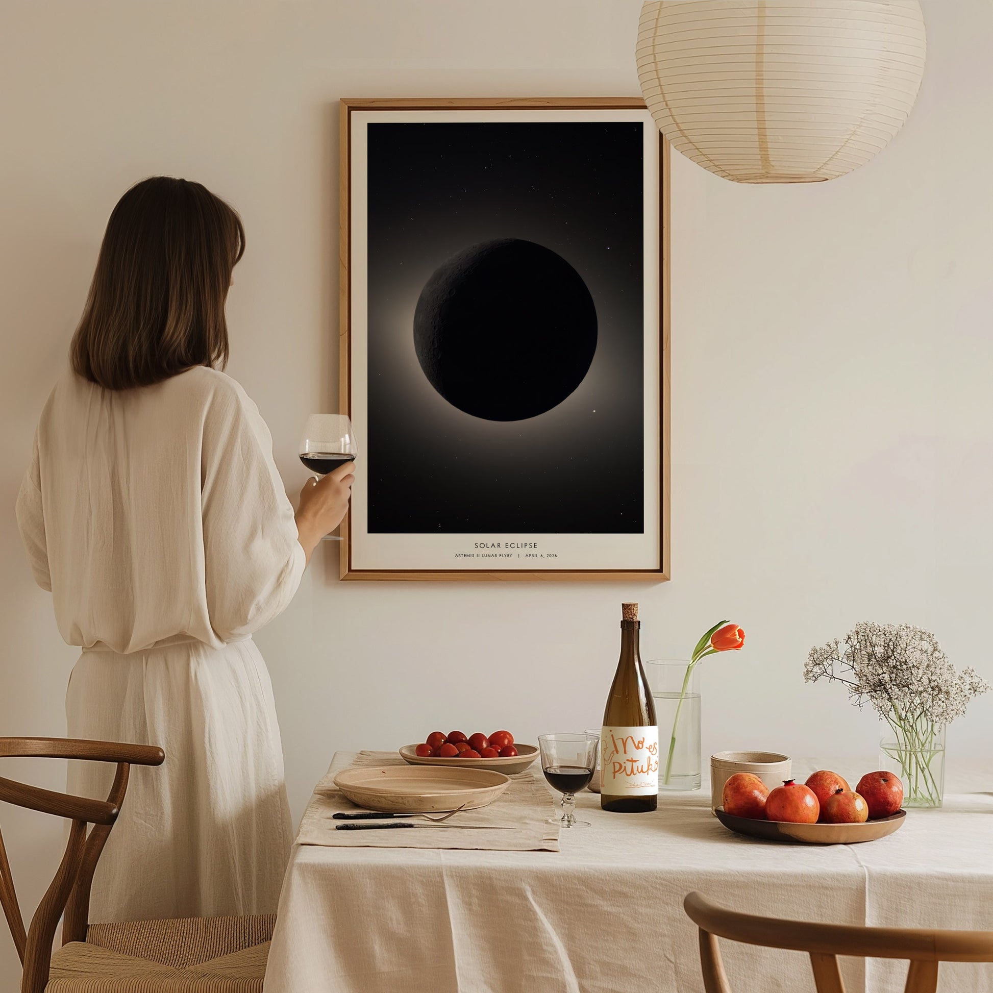 A woman in a white outfit holds a glass of wine while standing beside a dining table set with fruit and a bottle. On the wall, theres a framed poster depicting a solar eclipse, creating a serene, elegant atmosphere.