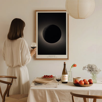 A woman in a white outfit holds a glass of wine while standing beside a dining table set with fruit and a bottle. On the wall, theres a framed poster depicting a solar eclipse, creating a serene, elegant atmosphere.