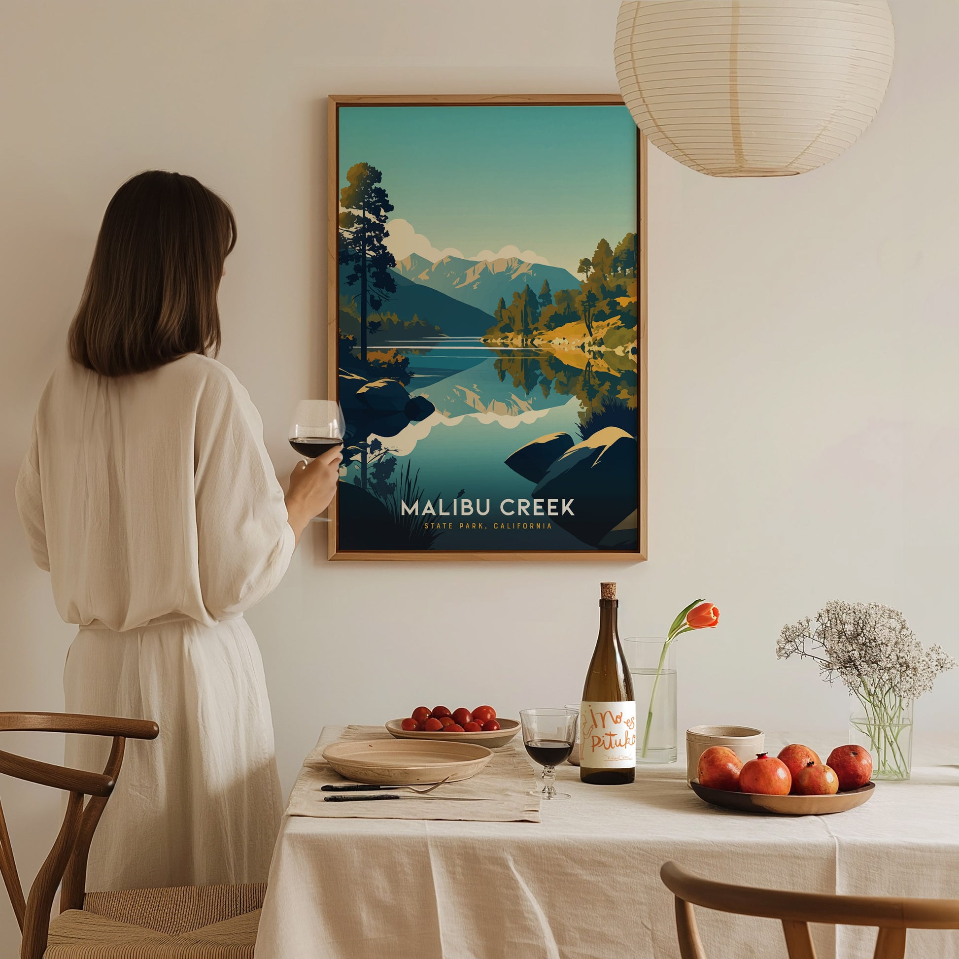 A woman in a white dress admires a colorful Malibu Creek State Park, California poster. A dining table with wine, fruit, and flowers is set in the foreground.
